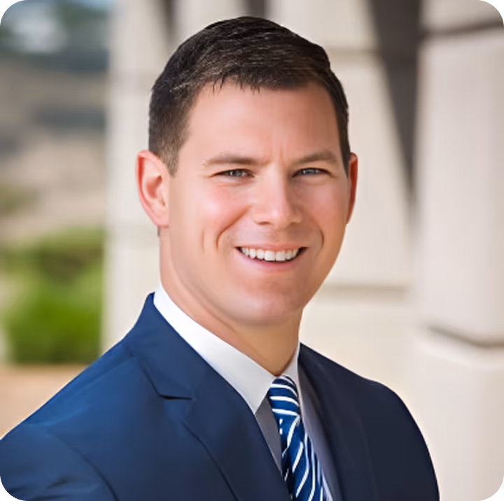 Professional headshot of smiling man in navy blue suit and striped tie standing outside a modern building