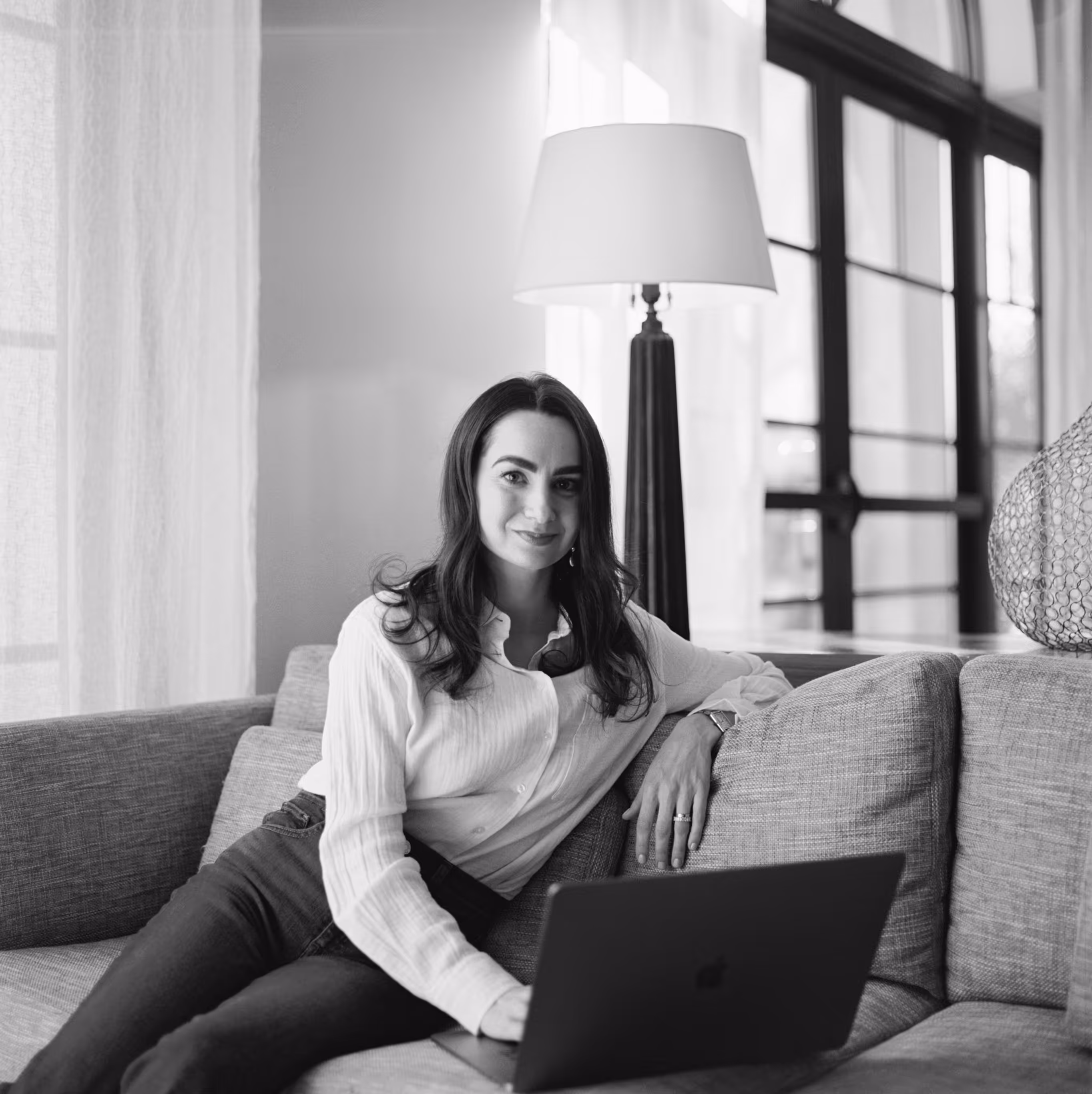Woman with dark hair sitting on a couch working on a laptop in a bright room.