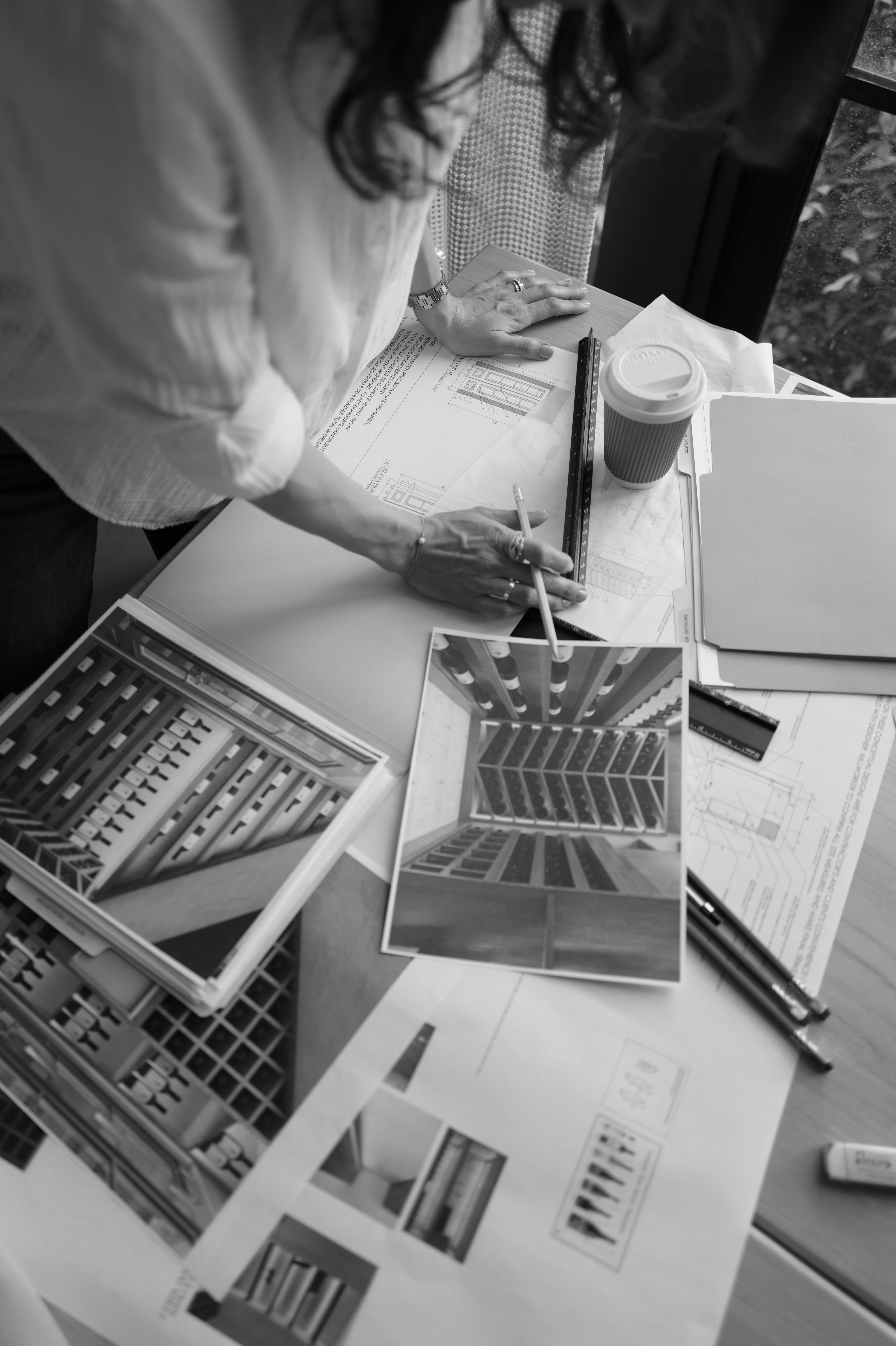Person working at a desk with architectural blueprints, design photos, a ruler, pens, and a takeout coffee cup.