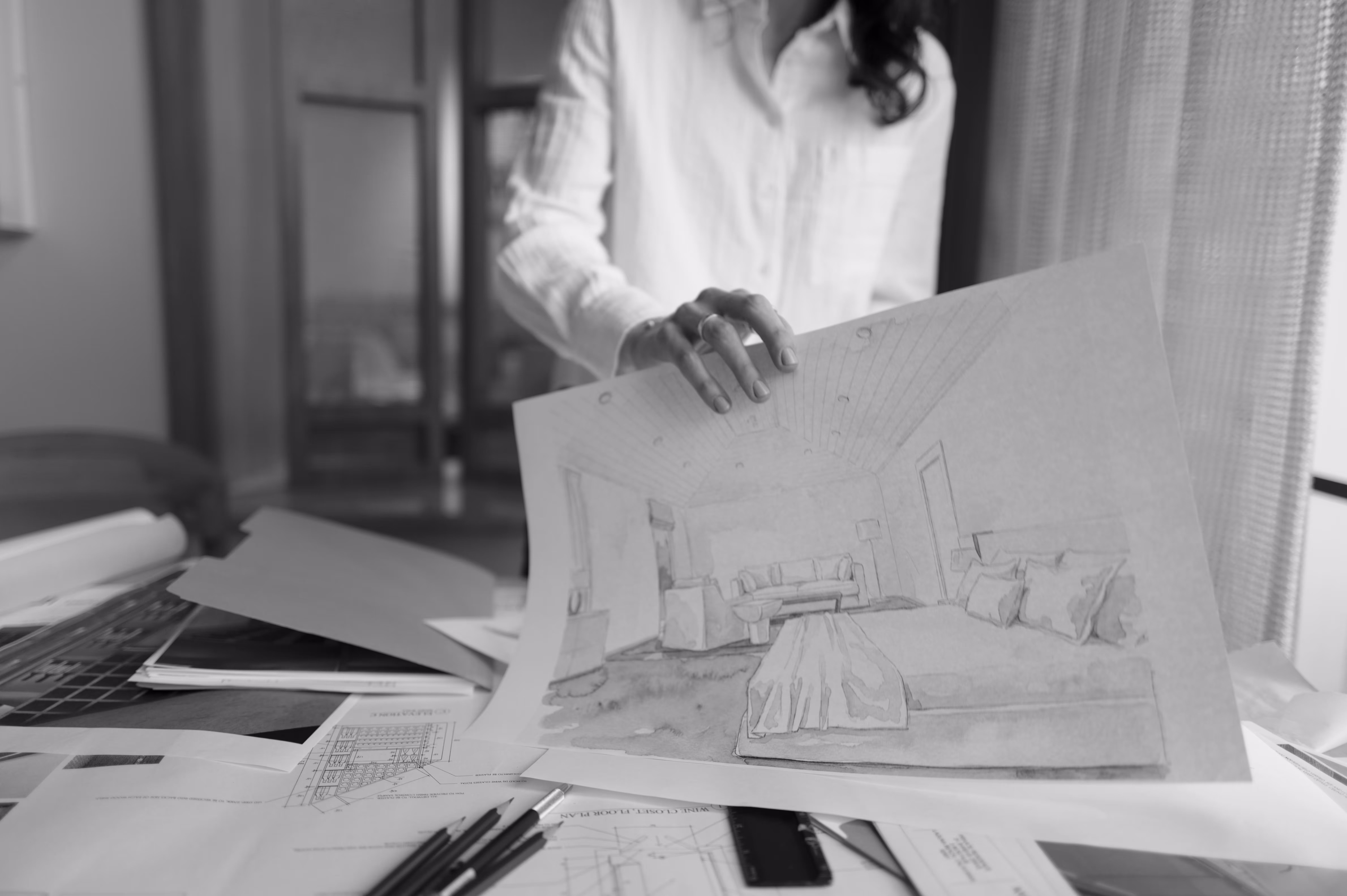 Person holding an architectural sketch of a bedroom interior over a cluttered desk with pencils and blueprints.