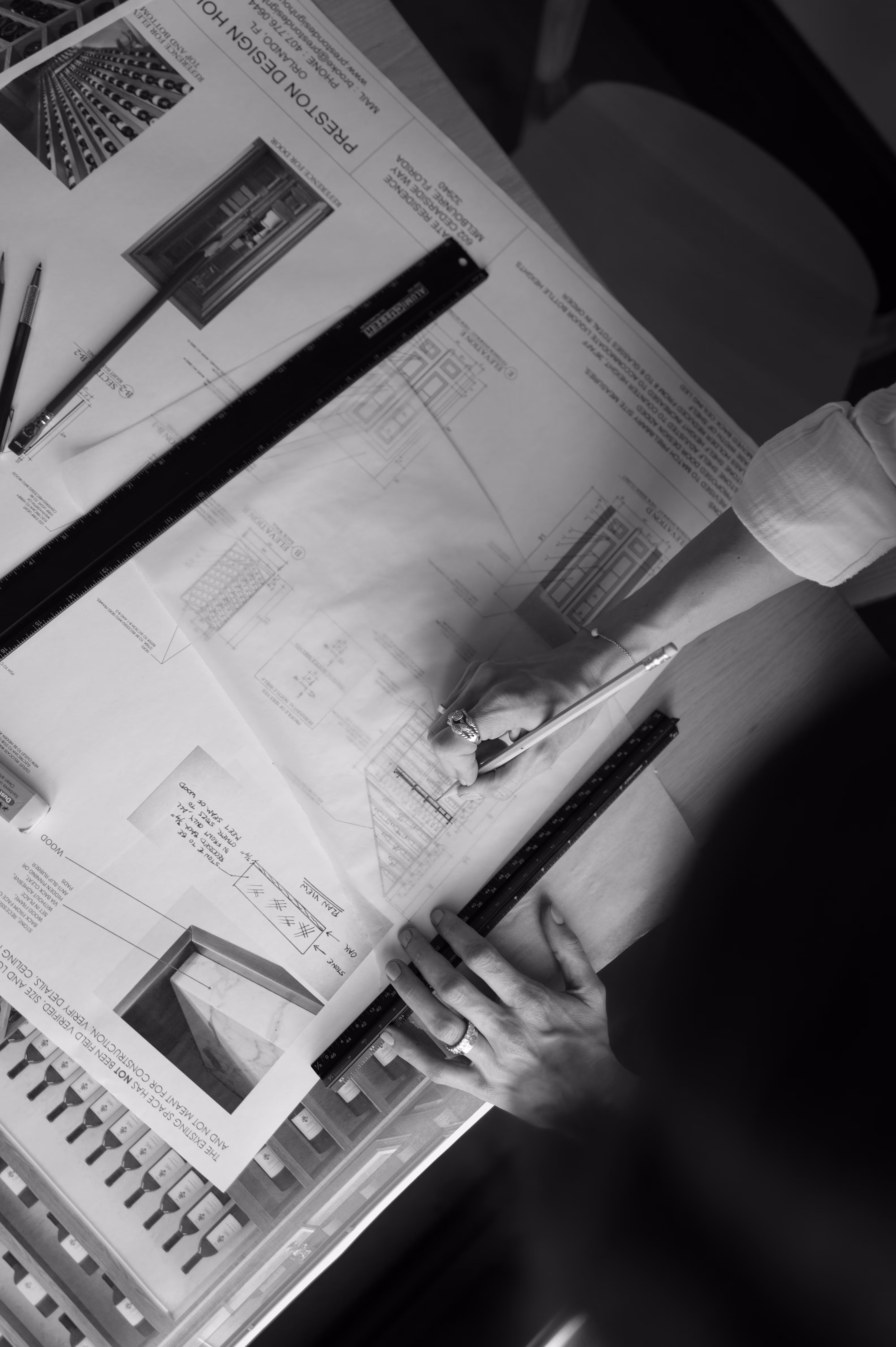 Person drawing architectural designs on paper with rulers and pencils on a wooden desk.