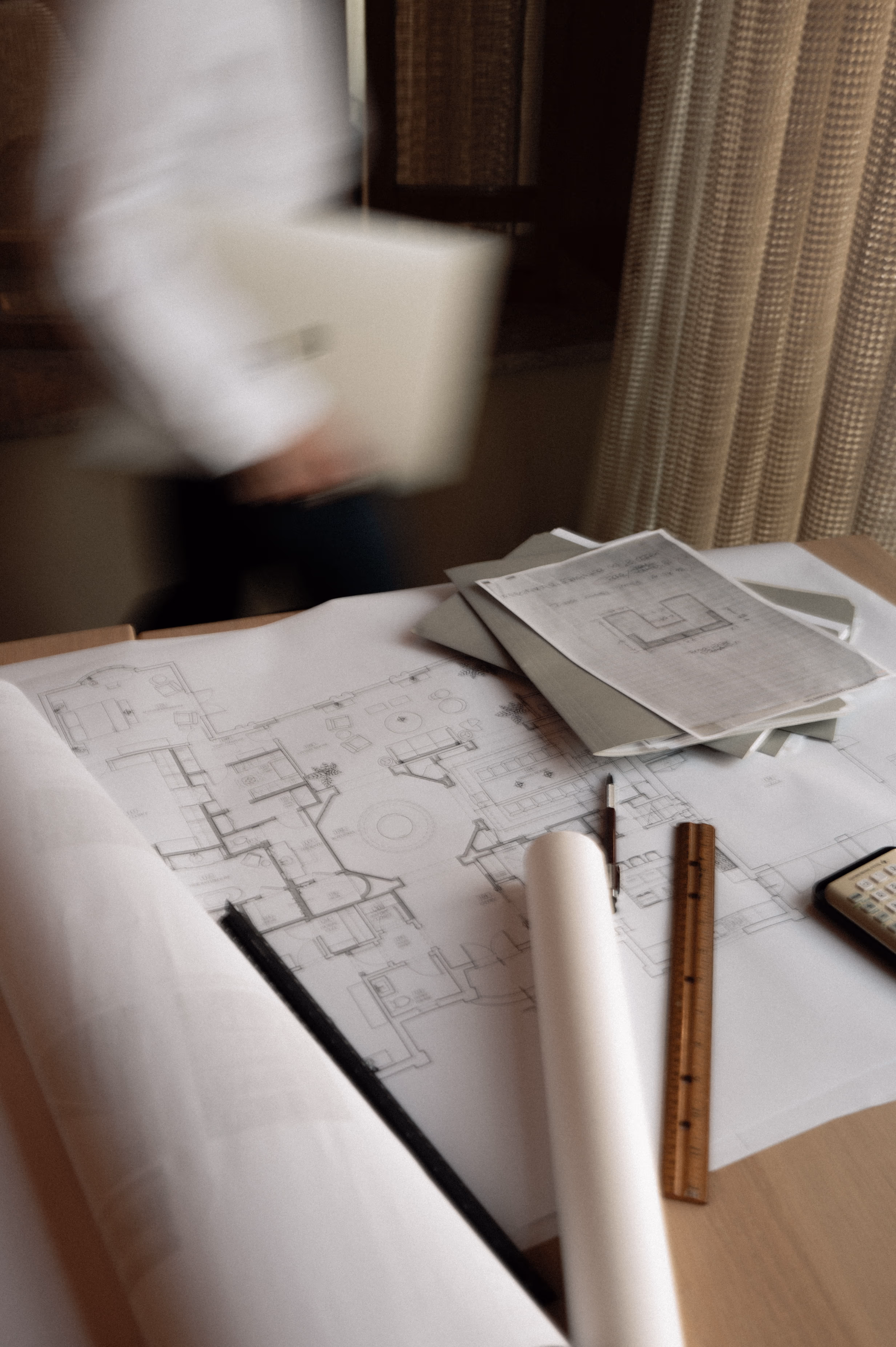 Architectural blueprints and drafting tools including a ruler and calculator on a wooden table with a blurred person holding a folder in the background.