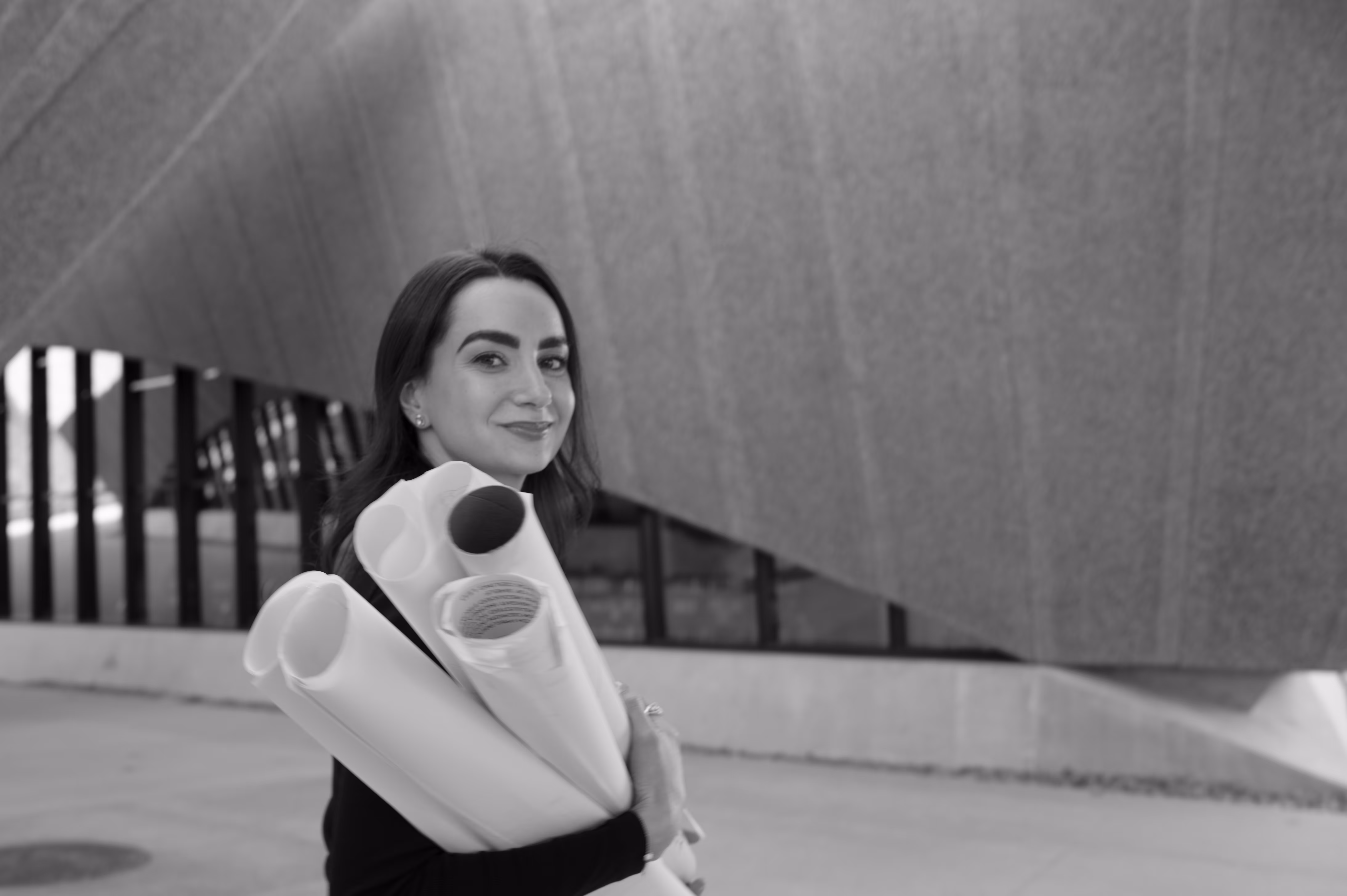 Smiling woman carrying rolled architectural blueprints outside a modern building.