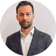 Portrait of a man with short dark hair and beard wearing a dark blazer and white shirt against a light gray background.