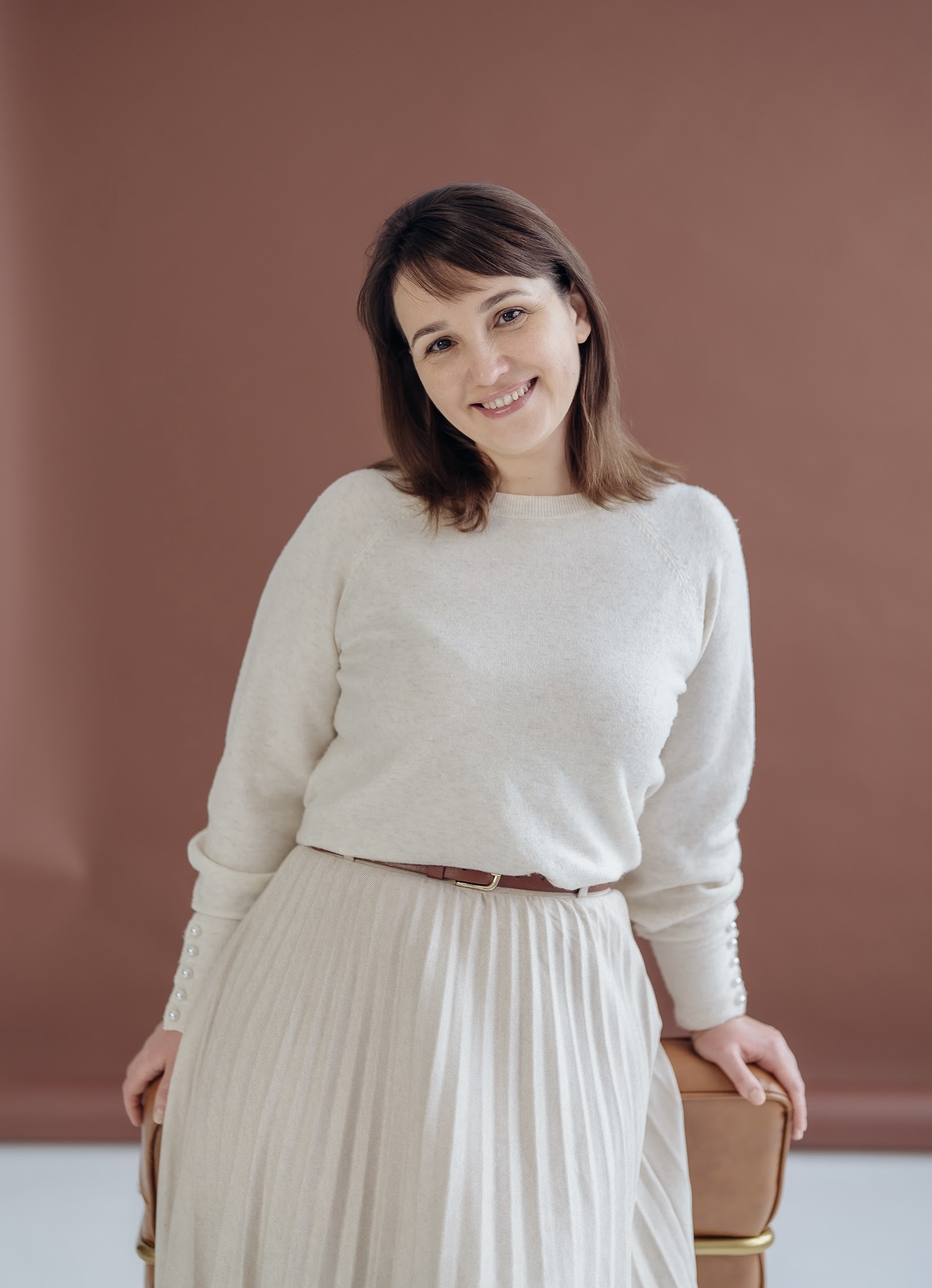 Smiling woman with brown hair wearing a white sweater and pleated skirt leaning on a brown chair against a muted brown background.