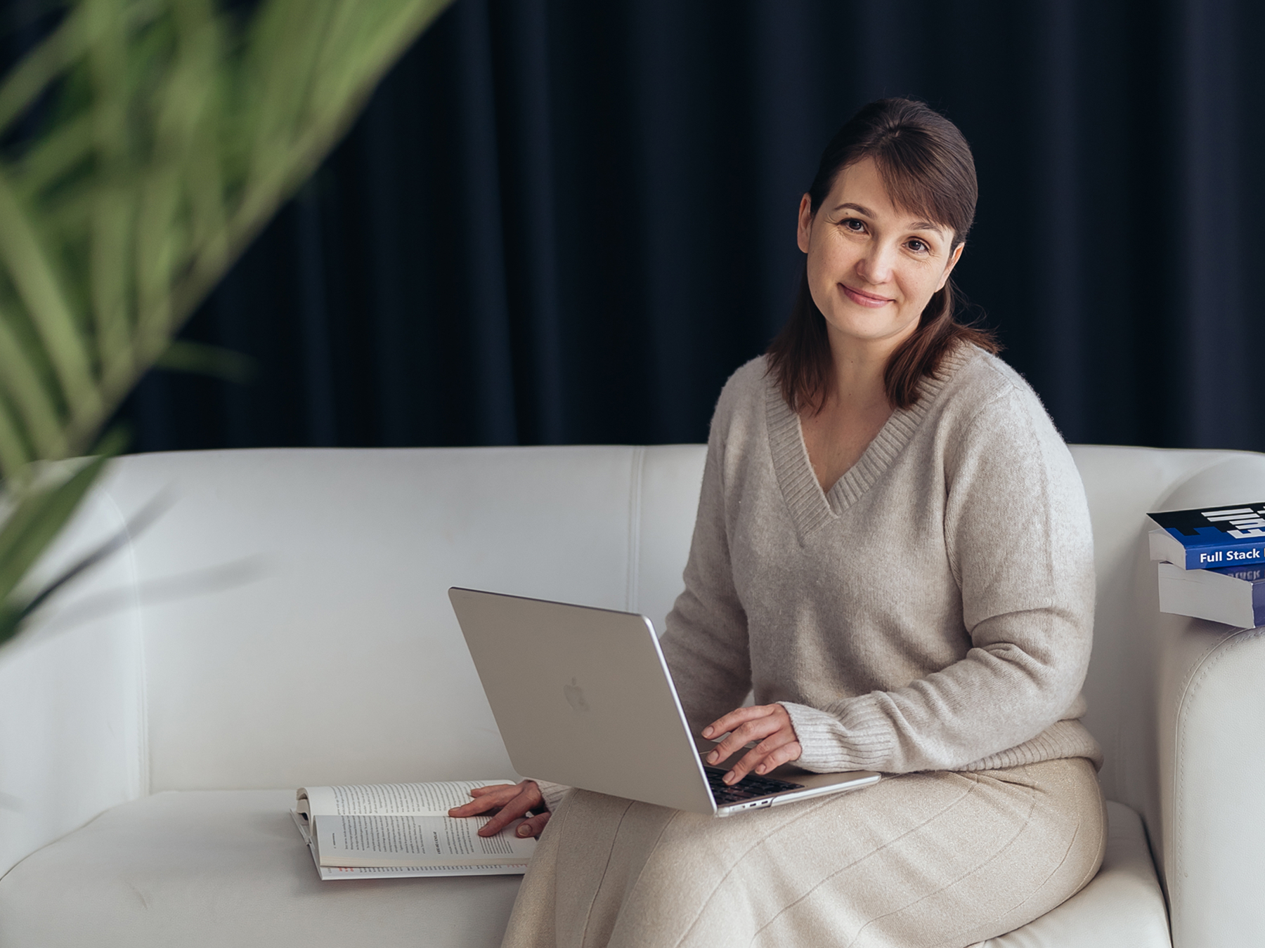 A woman in a beige sweater and skirt sitting on a white couch using a silver laptop, with an open book on the couch beside her and a stack of books nearby.