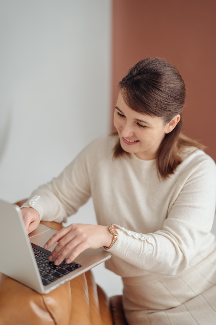 Smiling woman with brown hair in a beige sweater typing on a laptop while sitting on a chair.