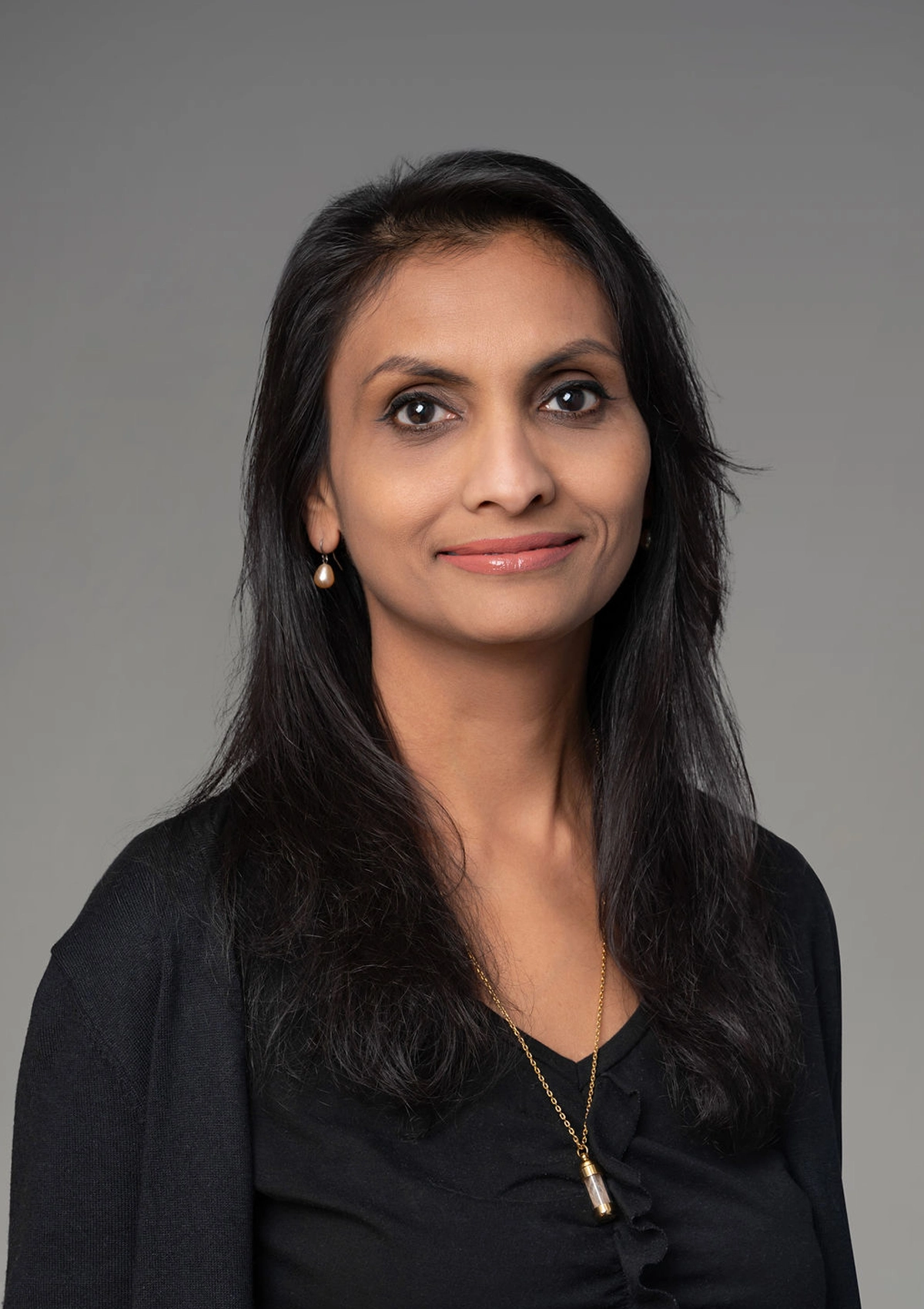 Woman with long black hair wearing black shirt and gold necklace, neutral expression against gray background