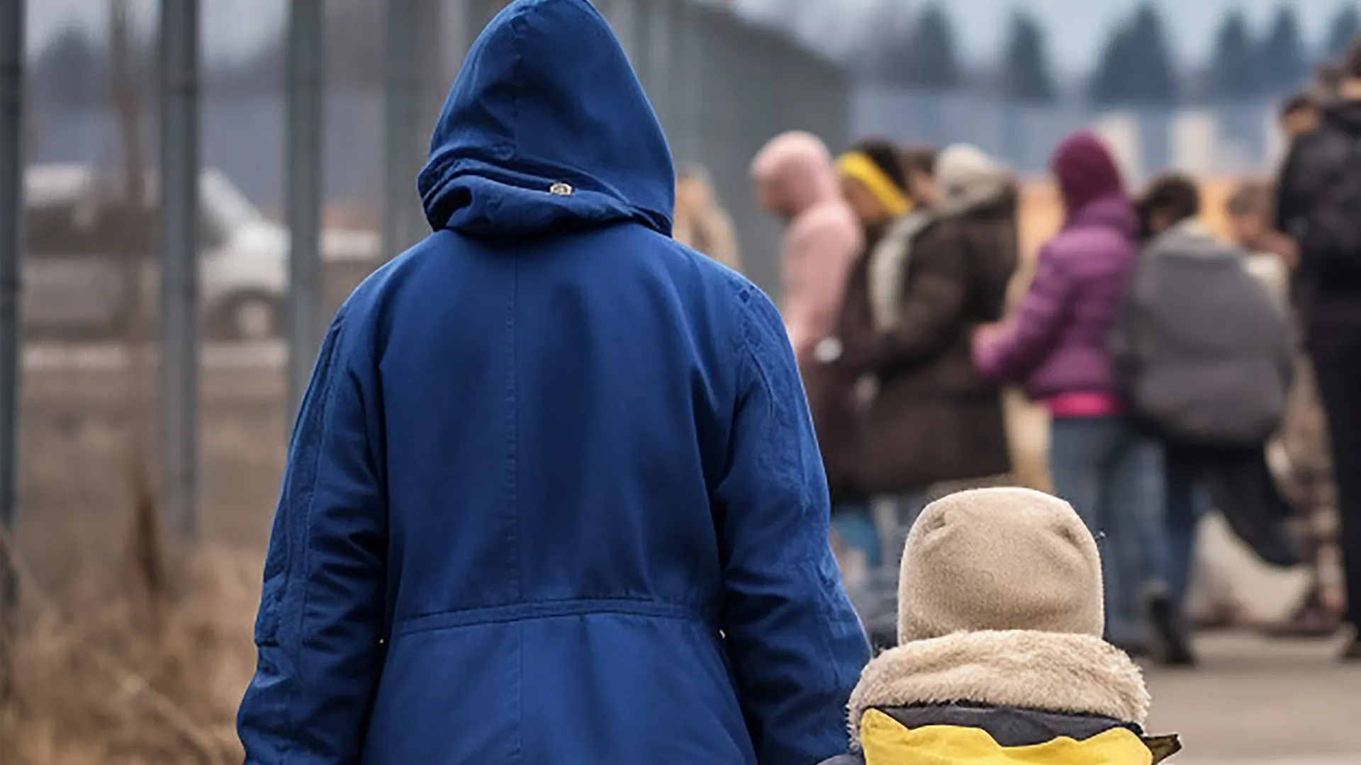 Child in blue jacket faces blurred crowd at refugee camp with fencing behind