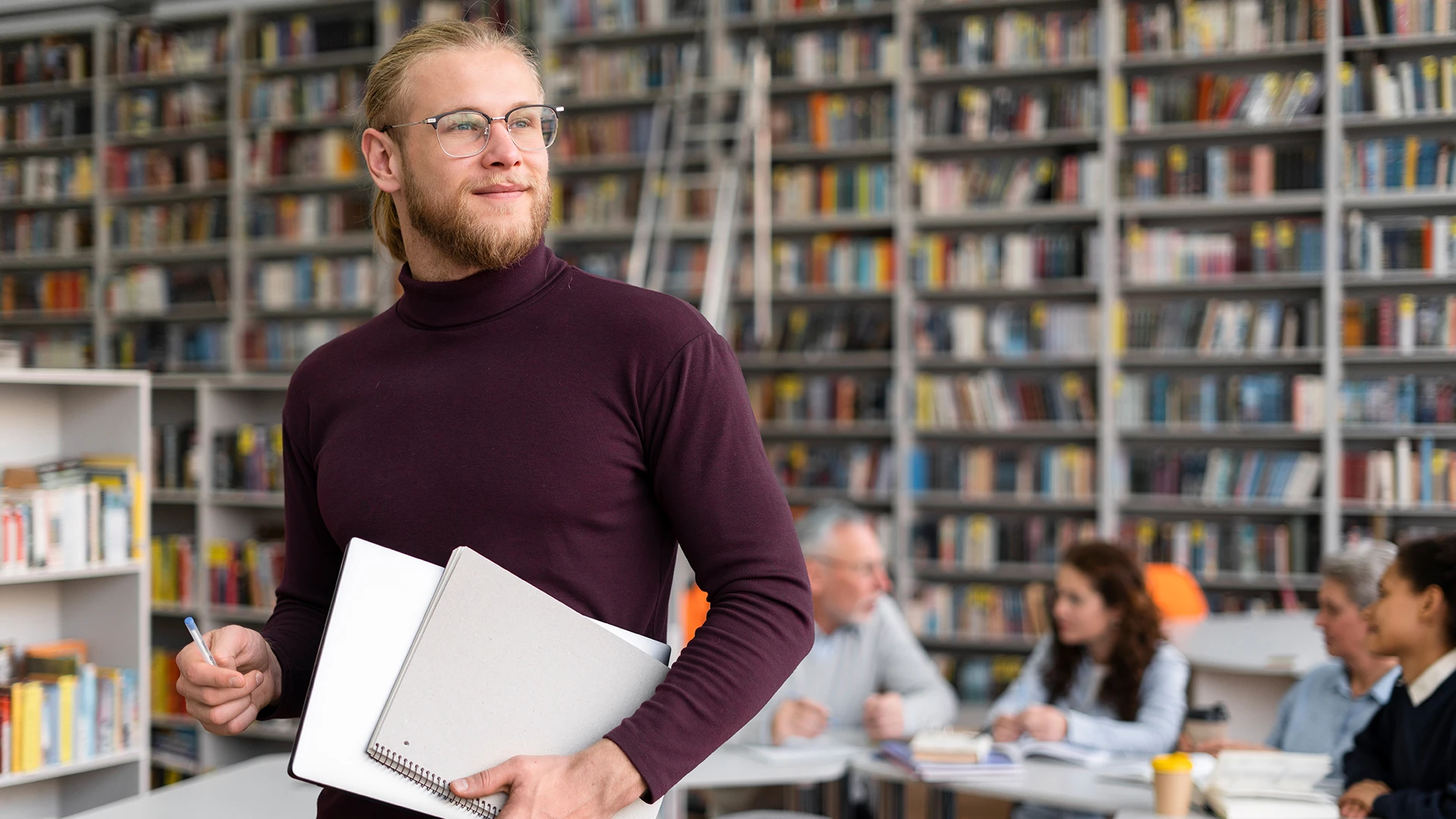 Man with notebook stands in library with bookshelves and seated colleagues in background.