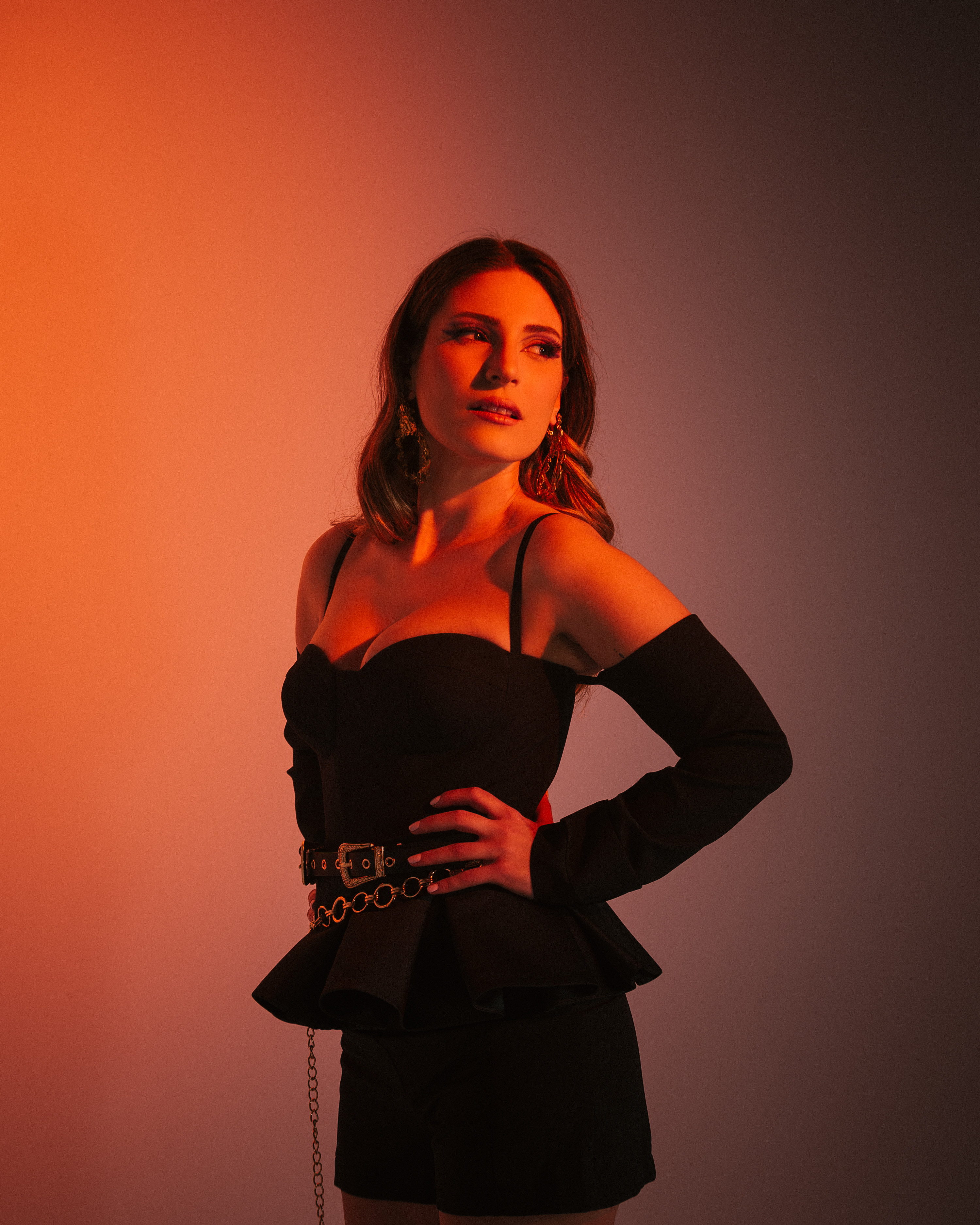 Woman in black off-shoulder dress with chain belt against orange gradient background