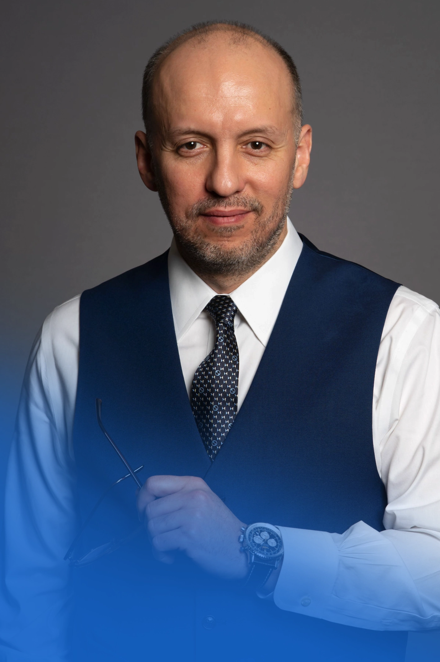 Professional portrait of a Remzi Kulen wearing navy vest, white shirt, and dark tie against blue background.