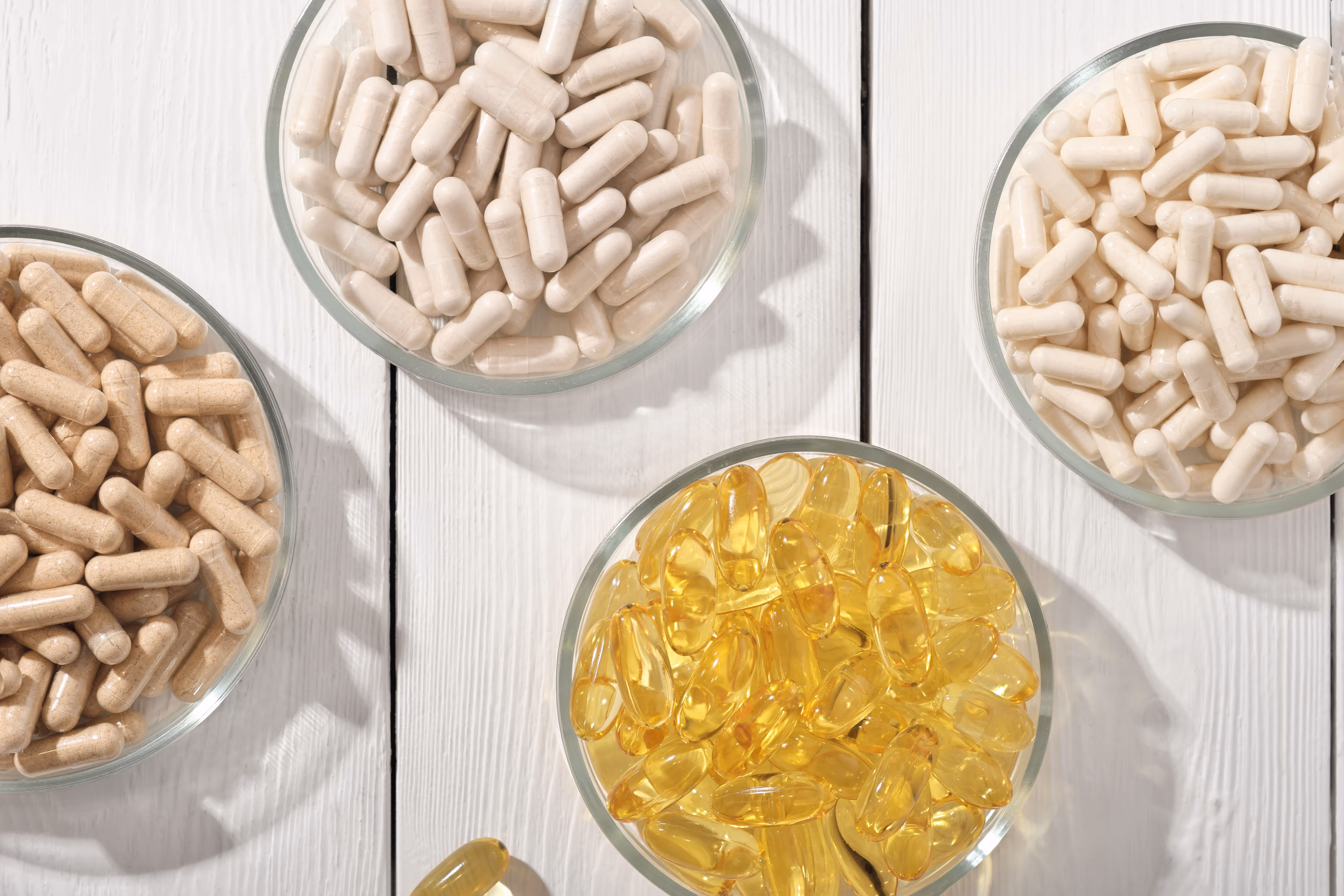 Quatre coupelles en verre contenant des capsules de suppléments diverses sur un fond de bois blanc.