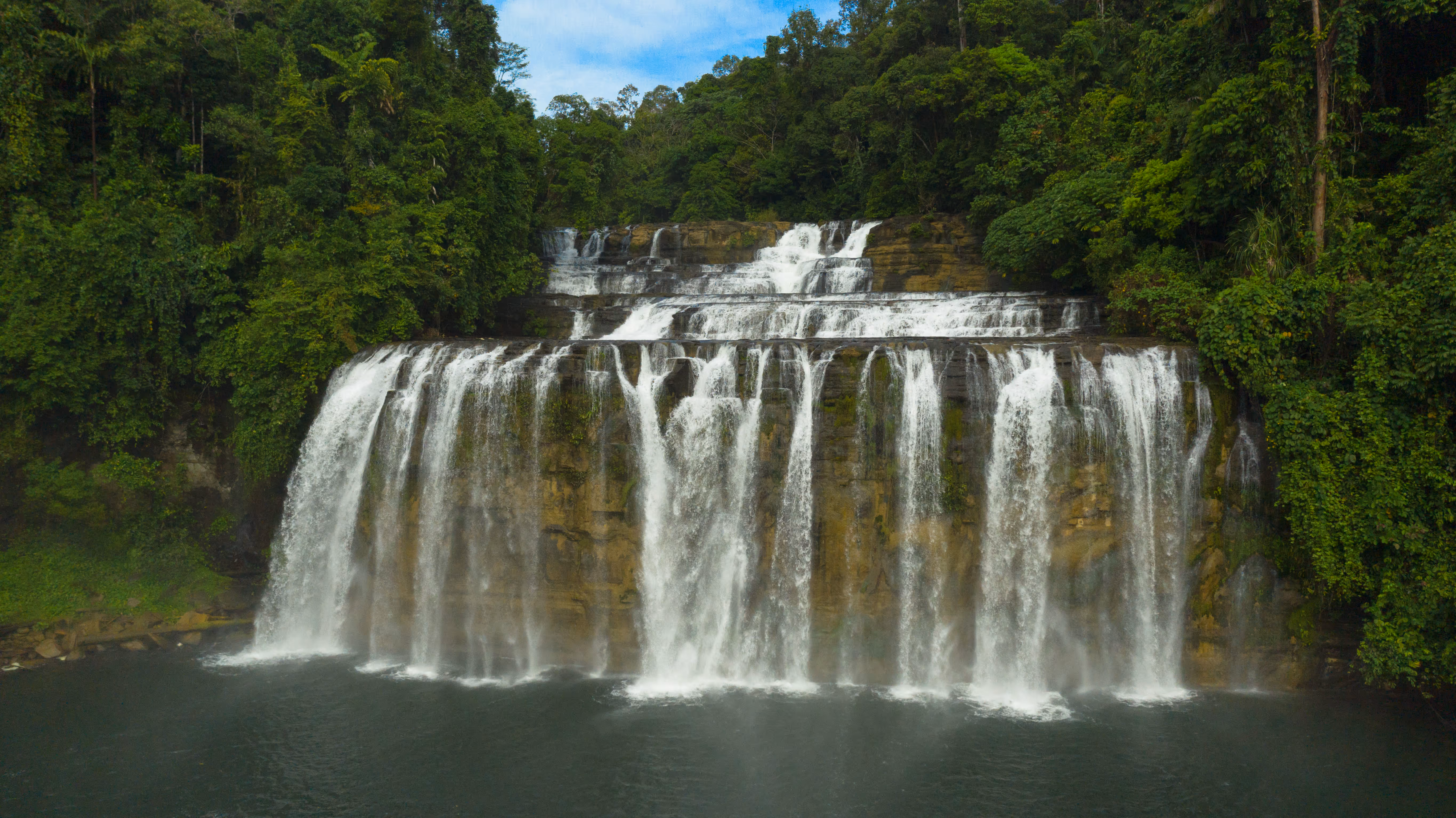 Cascade en terrasses larges entourée d'une végétation tropicale dense sous un ciel bleu.