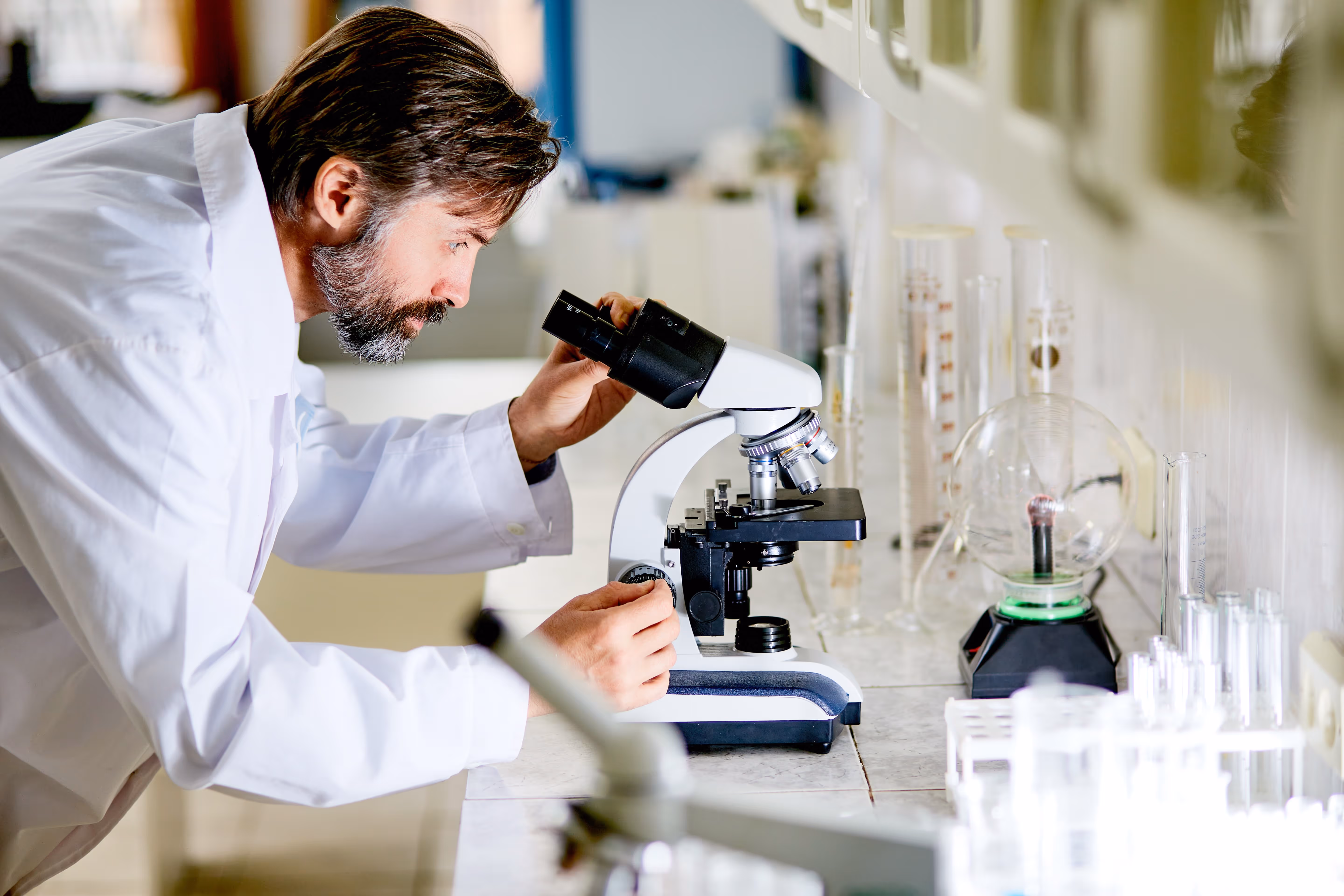 Un scientifique dans un laboratoire, observant à travers un microscope blanc avec divers tubes à essai et instruments en arrière-plan.