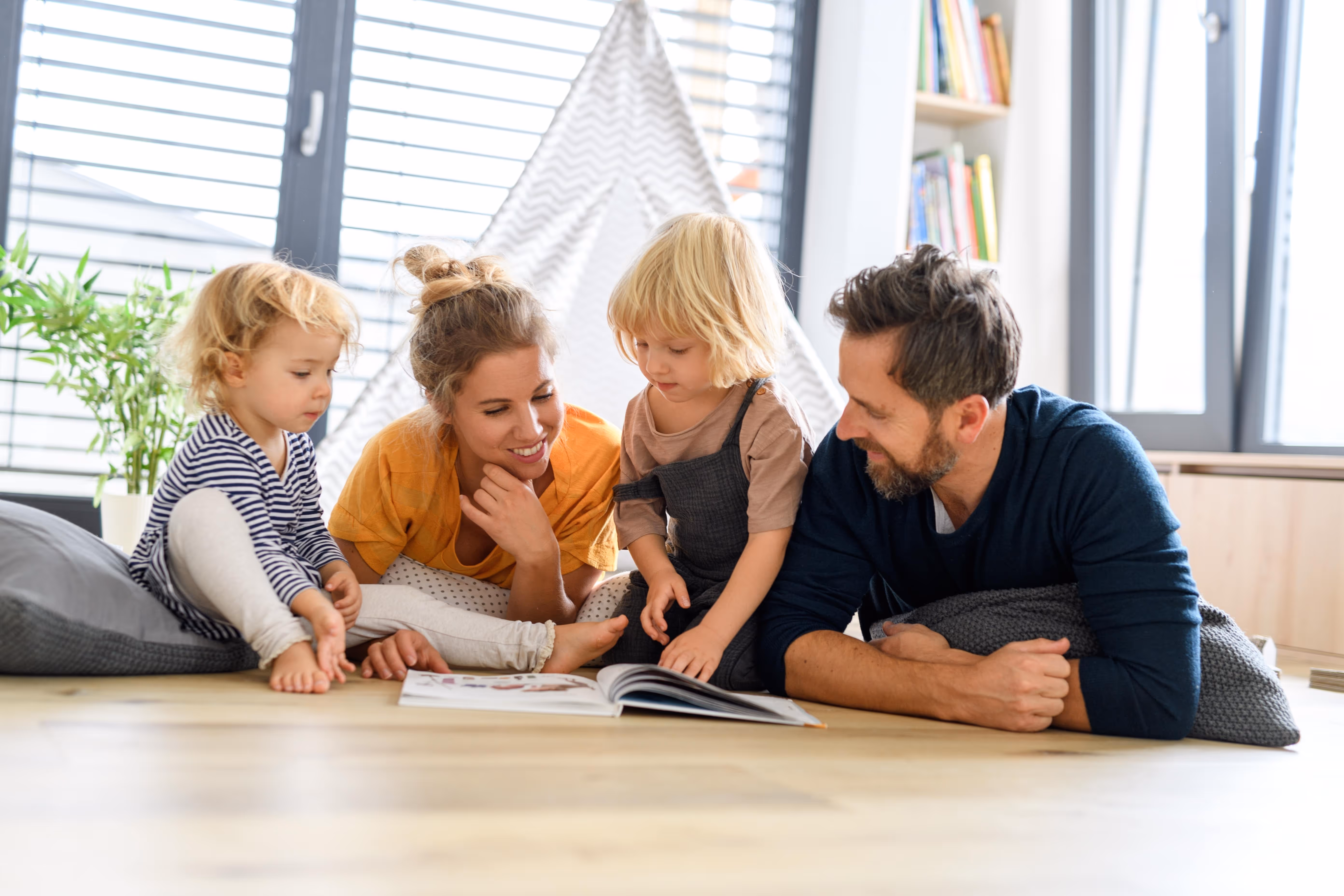 Une famille de quatre personnes, deux adultes et deux jeunes enfants, lisant un livre ensemble sur le sol dans un salon lumineux.