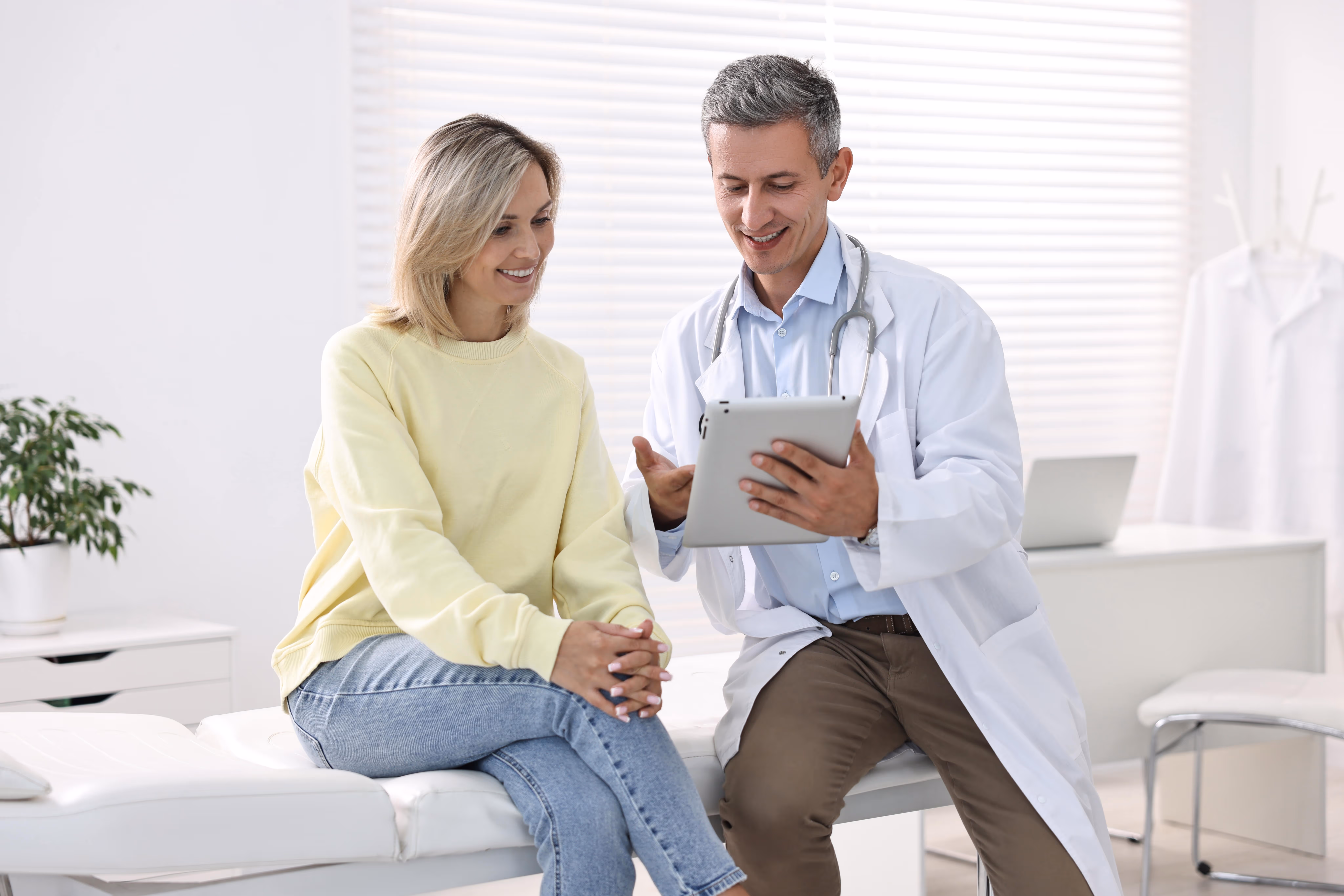 Un médecin souriant avec une blouse blanche montre des informations sur une tablette à une patiente assise qui écoute attentivement dans un cabinet médical lumineux.