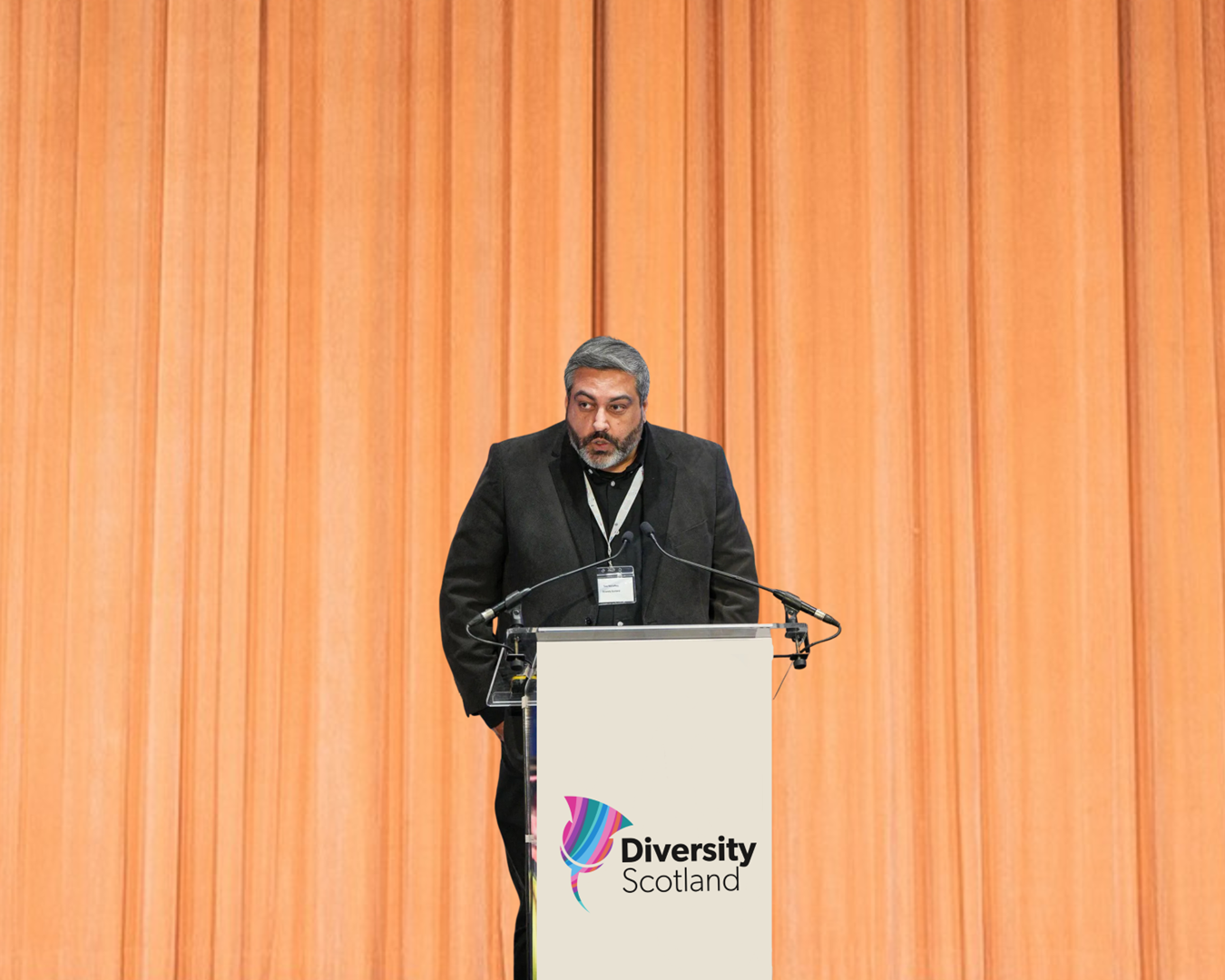 Tony McCaffery stands at a lectern on stage, speaking into a microphone. The lectern displays the Diversity Scotland logo, and the stage backdrop is a set of warm orange curtains.