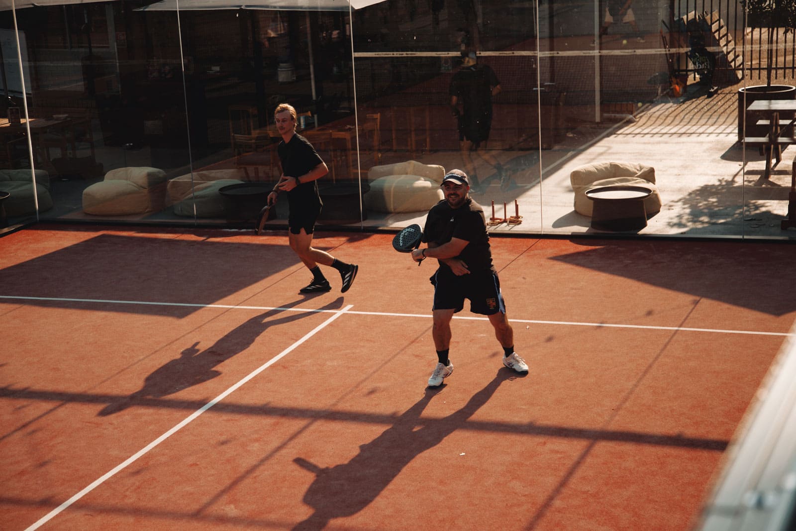 Two men playing paddle tennis on a red clay court enclosed by glass walls.