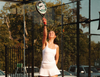 Smiling woman in white sportswear playing paddle tennis on an outdoor court, reaching up with a paddle.