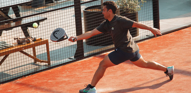 Man lunging to hit a ball with a paddle on an orange clay padel court.