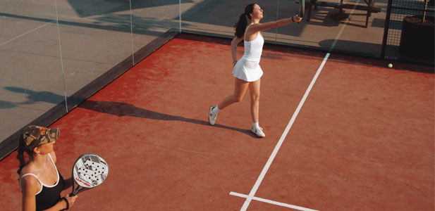 Two women playing paddle tennis on a clay court, one about to hit the ball and the other waiting with a paddle racket.
