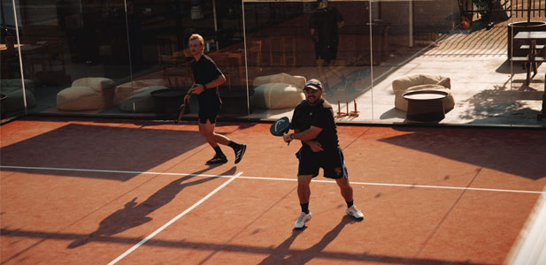 Two men playing paddle tennis on a sunlit clay court with glass walls and outdoor seating in the background.