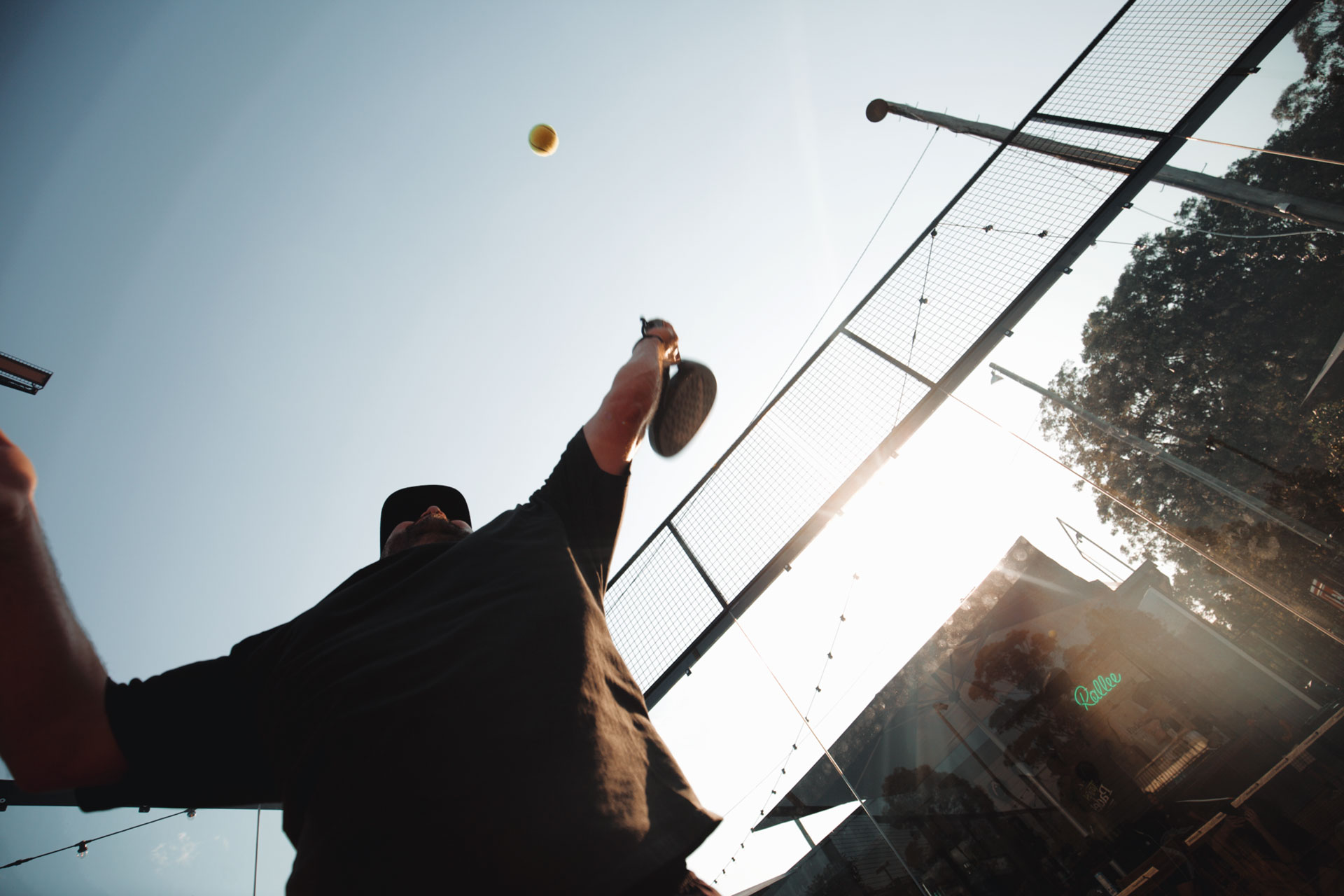 Low-angle view of a man playing pickleball, hitting a ball with a paddle on an outdoor court in sunlight.