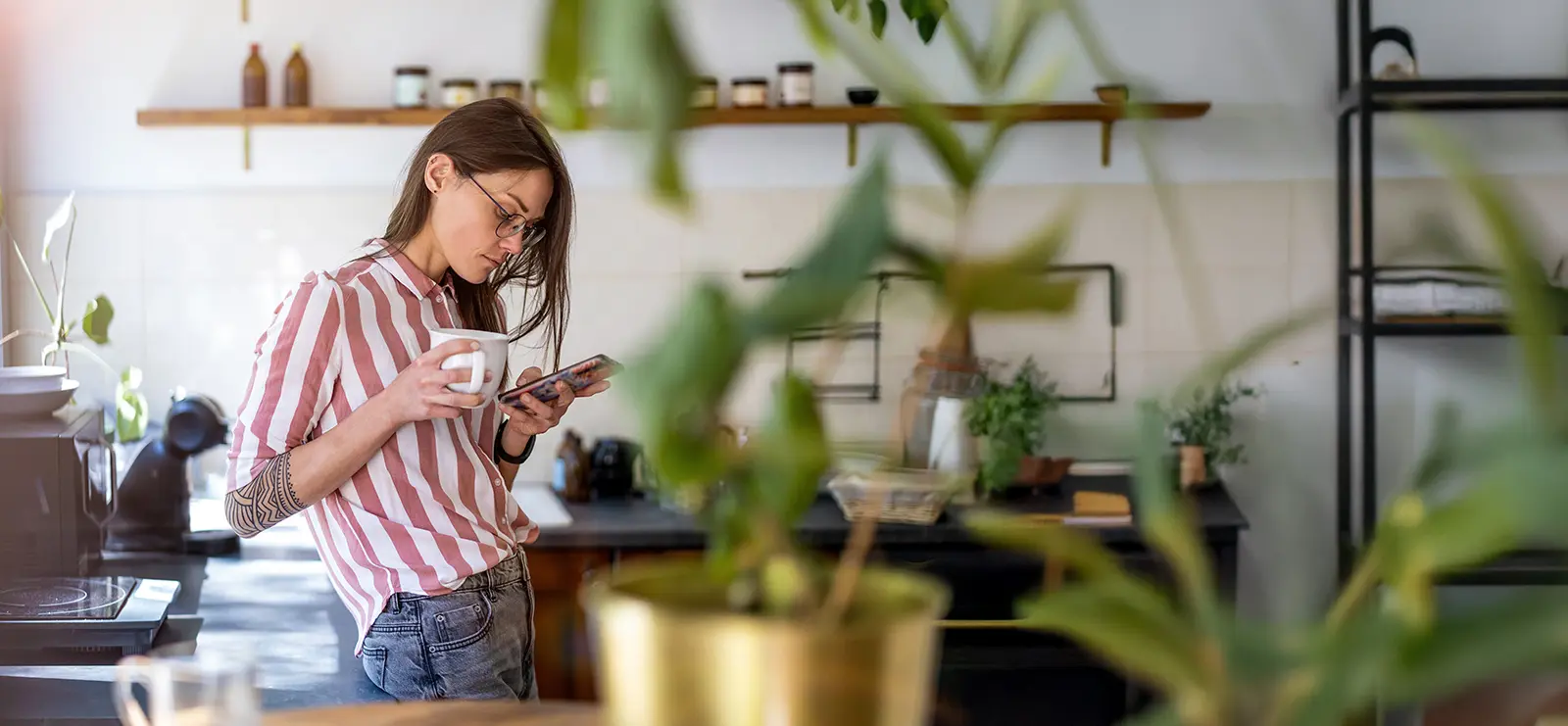 woman with tattoos looking down at her phone while drinking coffee in her kitchen