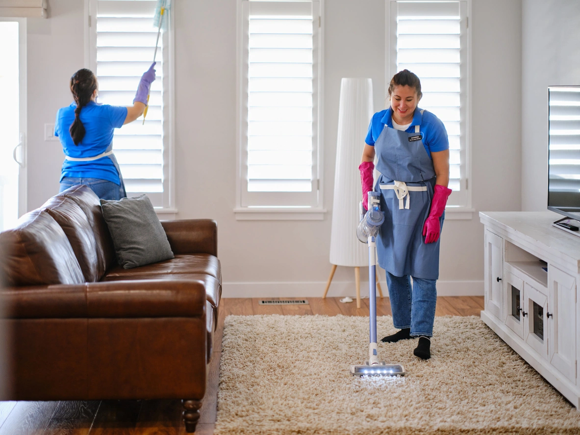 Two women vacuuming a living room, focused on cleaning the carpet and surrounding furniture.