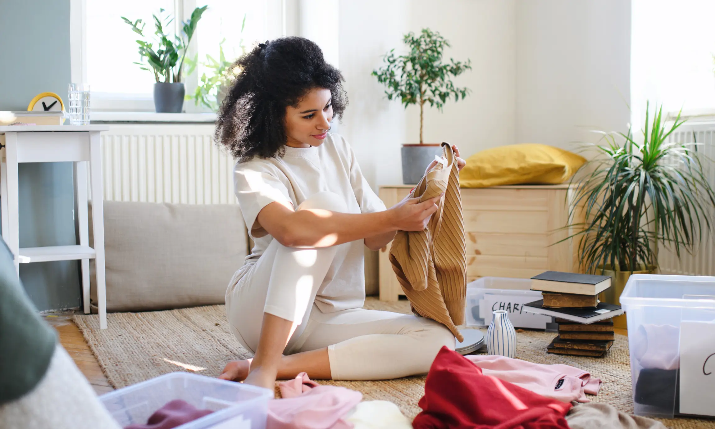 Woman sitting on the floor of a bright room, examining a piece of clothing. Around her are labeled plastic bins and scattered clothes, indicating she is sorting items for donation or organization. Books and indoor plants are visible in the background.