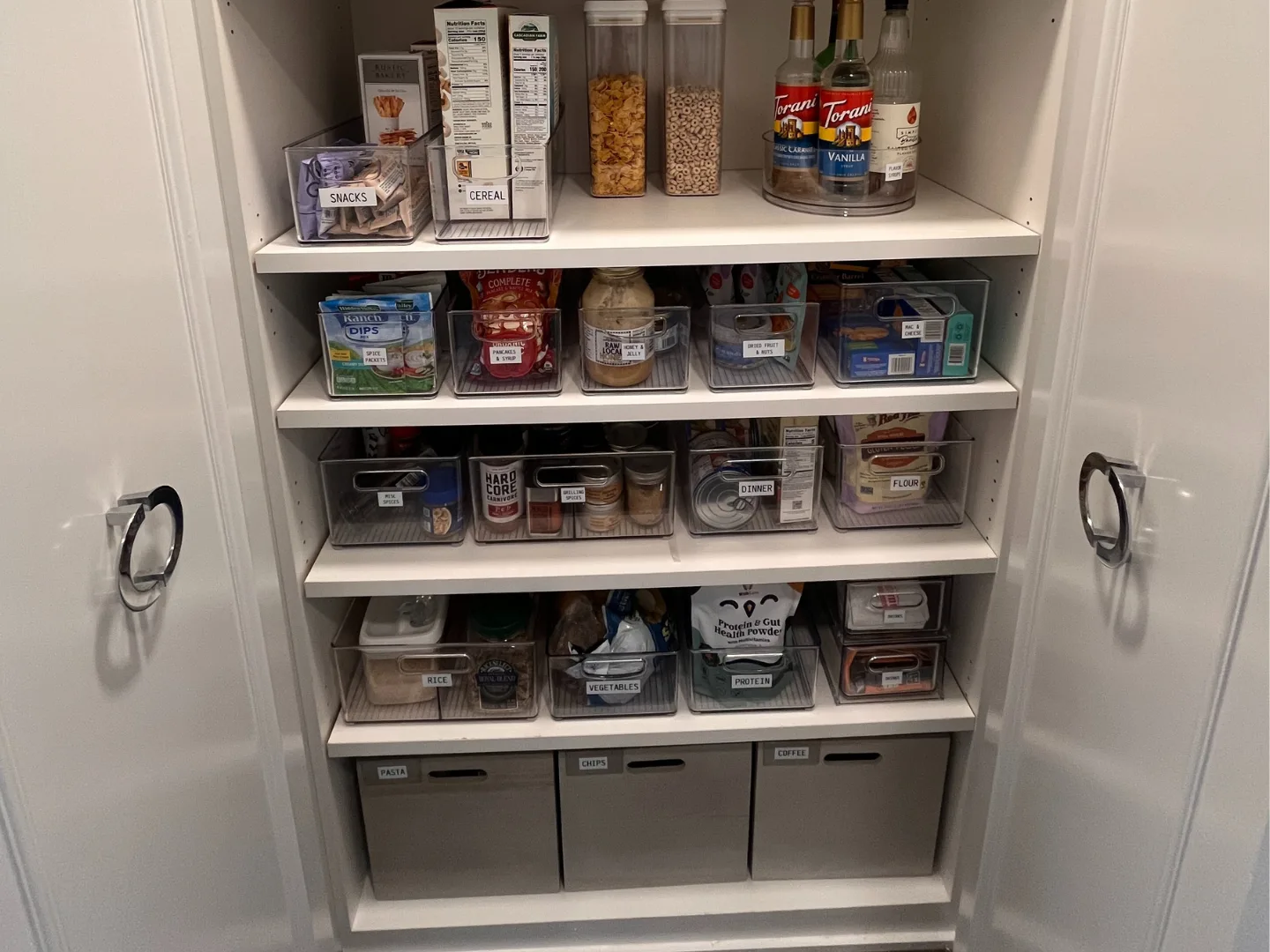 Open pantry cabinet with labeled clear bins on shelves containing snacks, cereal, sauces, grains, and baking ingredients, alongside turntables with bottles.