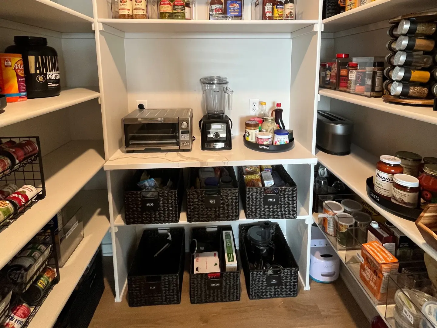 Walk-in pantry with black woven bins labeled by category, countertop with blender and toaster oven, and shelves stocked with spices, sauces, and canned goods.
