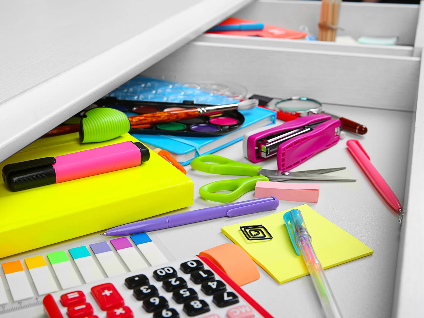 Open desk drawer with scattered office supplies including highlighters, scissors, sticky notes, calculator, paper clips, and a stapler in disarray.