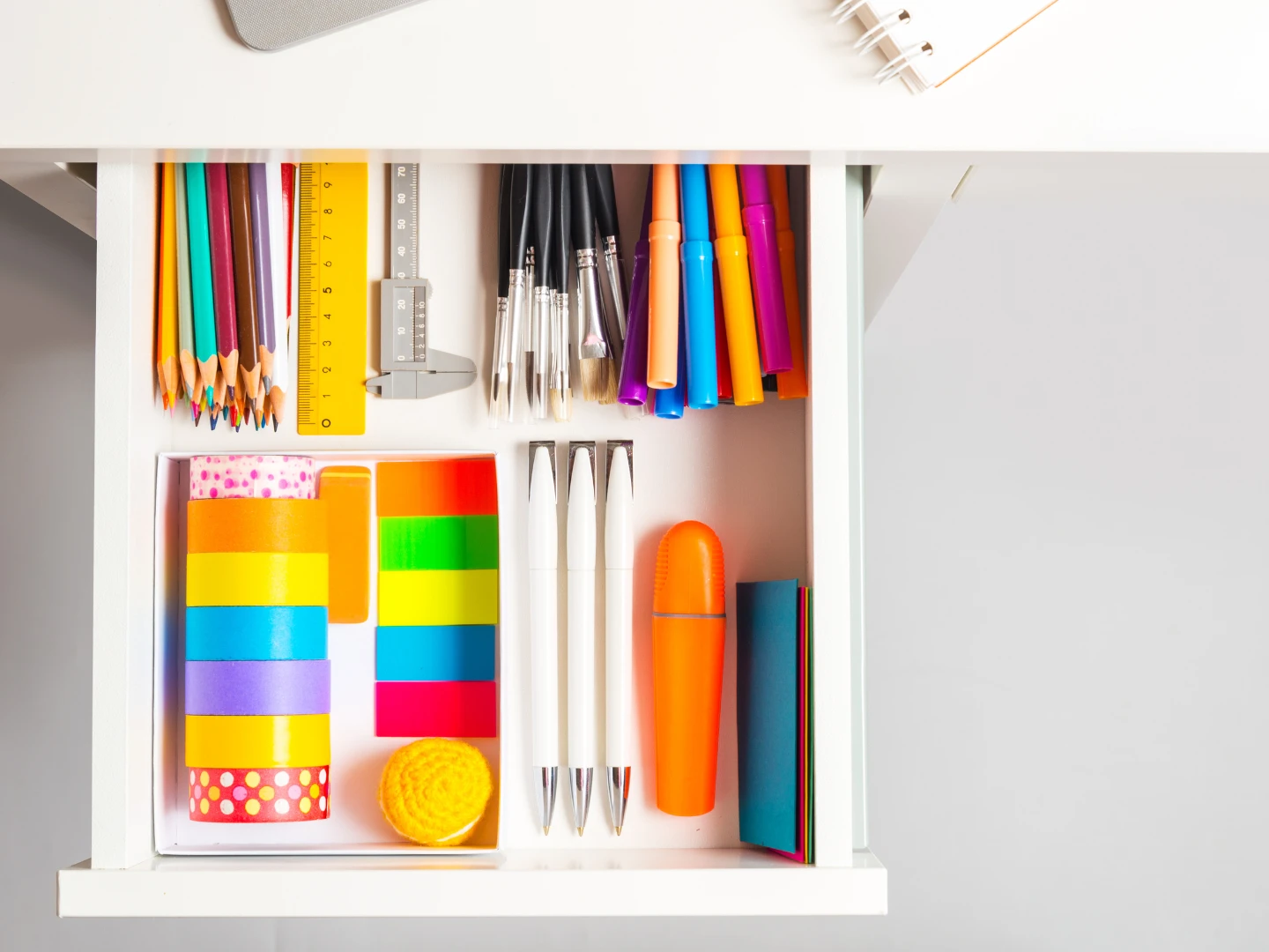 Overhead view of an organized drawer with grouped office supplies including colored pencils, pens, rulers, markers, washi tape, sticky notes, and a yellow pom-pom ball.
