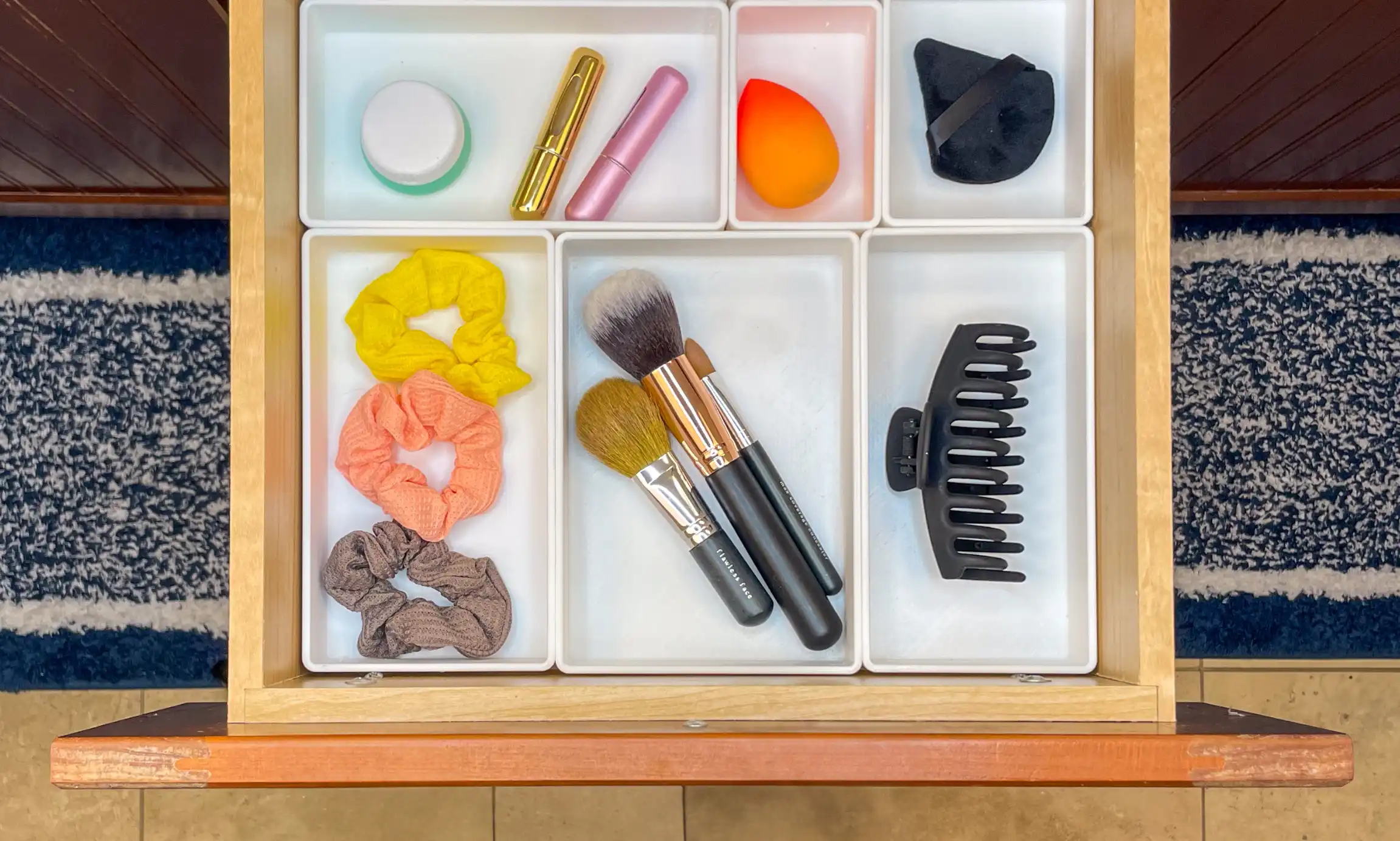 Open wooden drawer with white drawer organizers holding makeup items, three scrunchies, three makeup brushes, a black hair clip, a jar, two lipsticks, a makeup sponge, and a black makeup applicator puff.