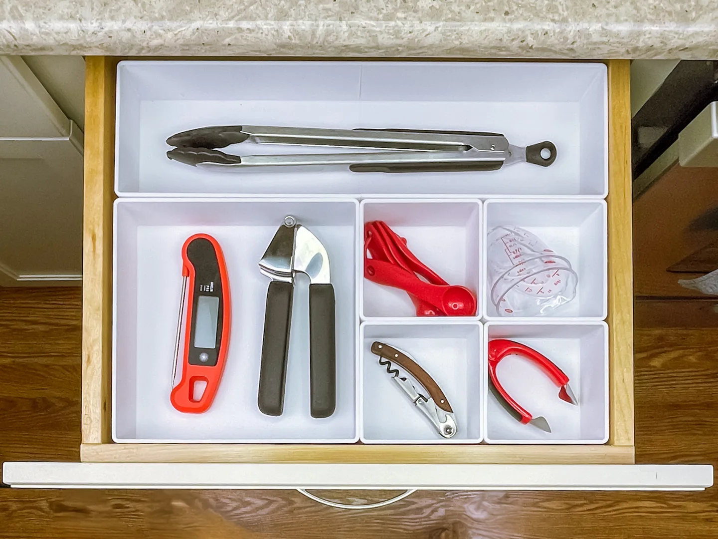 Overhead view of a kitchen drawer with white drawer organizers holding metal tongs, a digital thermometer, garlic press, red measuring spoons, clear measuring cup, corkscrew, and red wine foil cutter.
