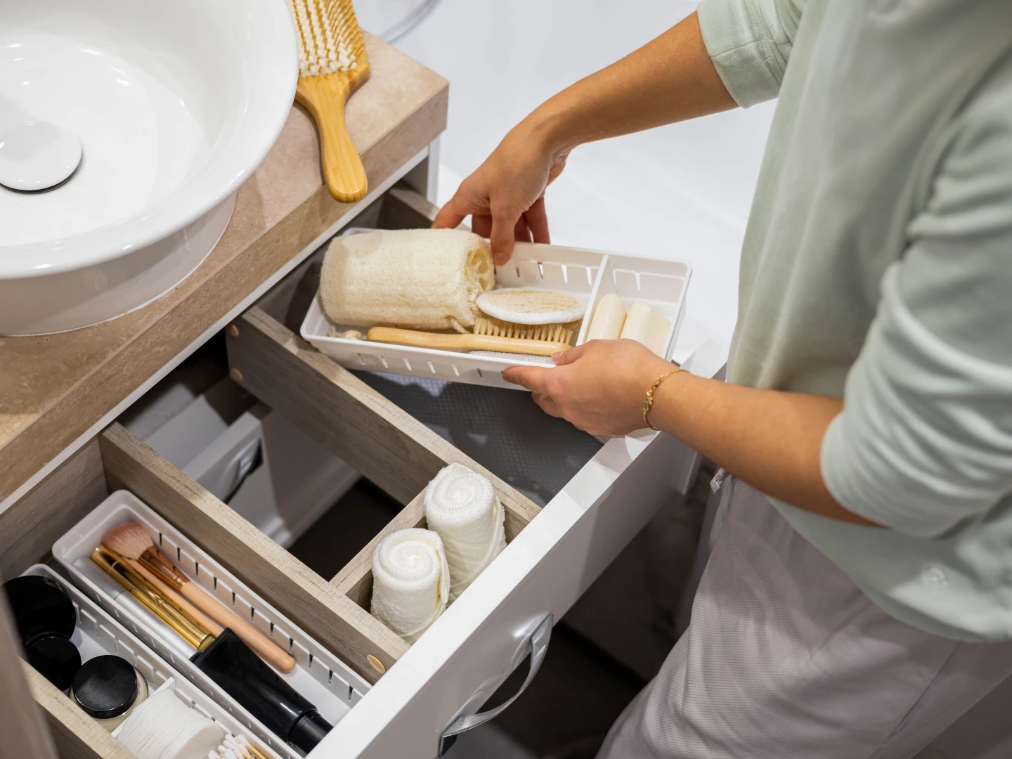 Person placing a plastic tray with rolled towels, soaps, and brushes into a bathroom drawer with other organized items.