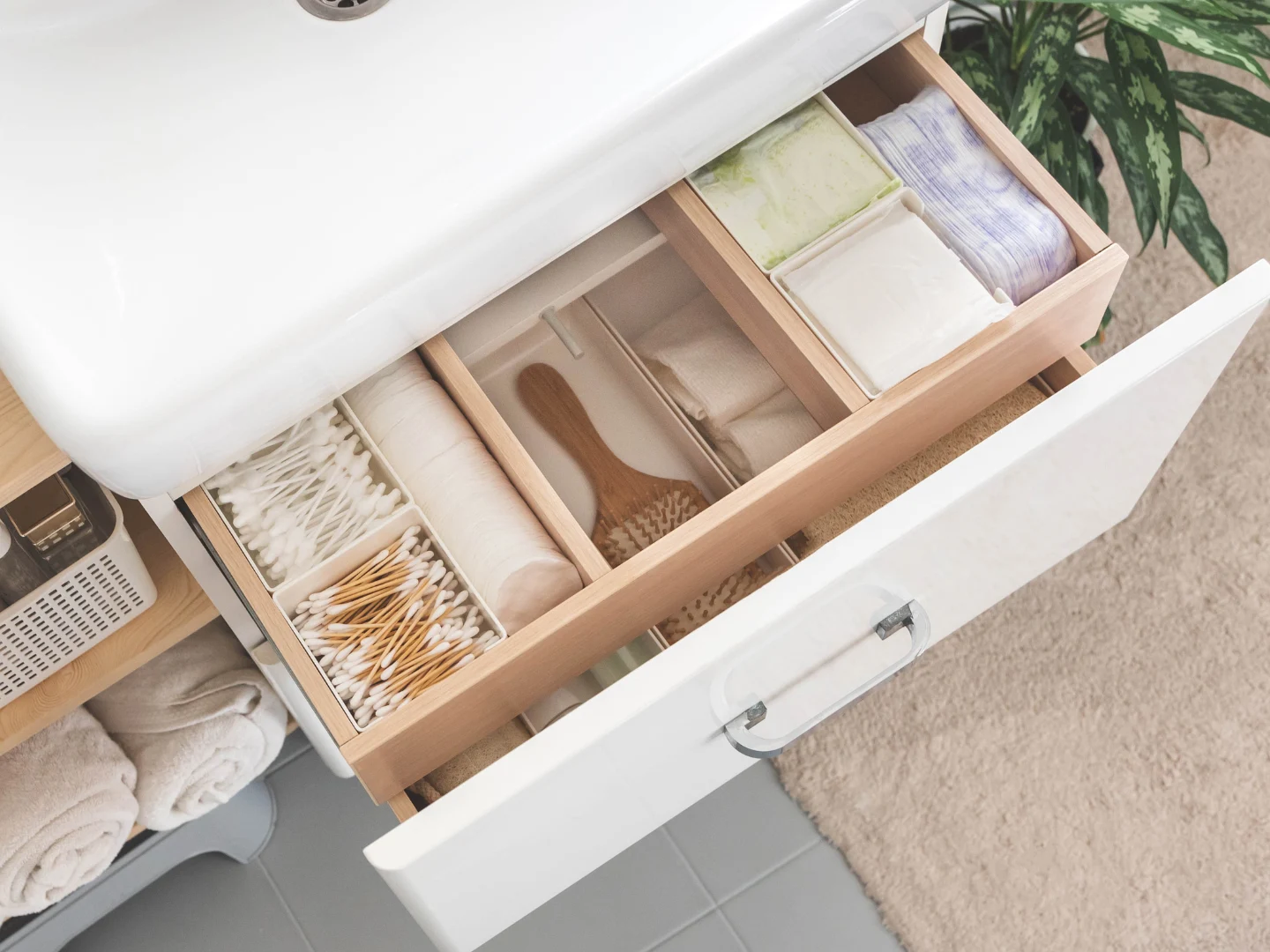 Open bathroom drawer with wooden dividers containing cotton swabs, soap bars, folded cloths, and a wooden brush.