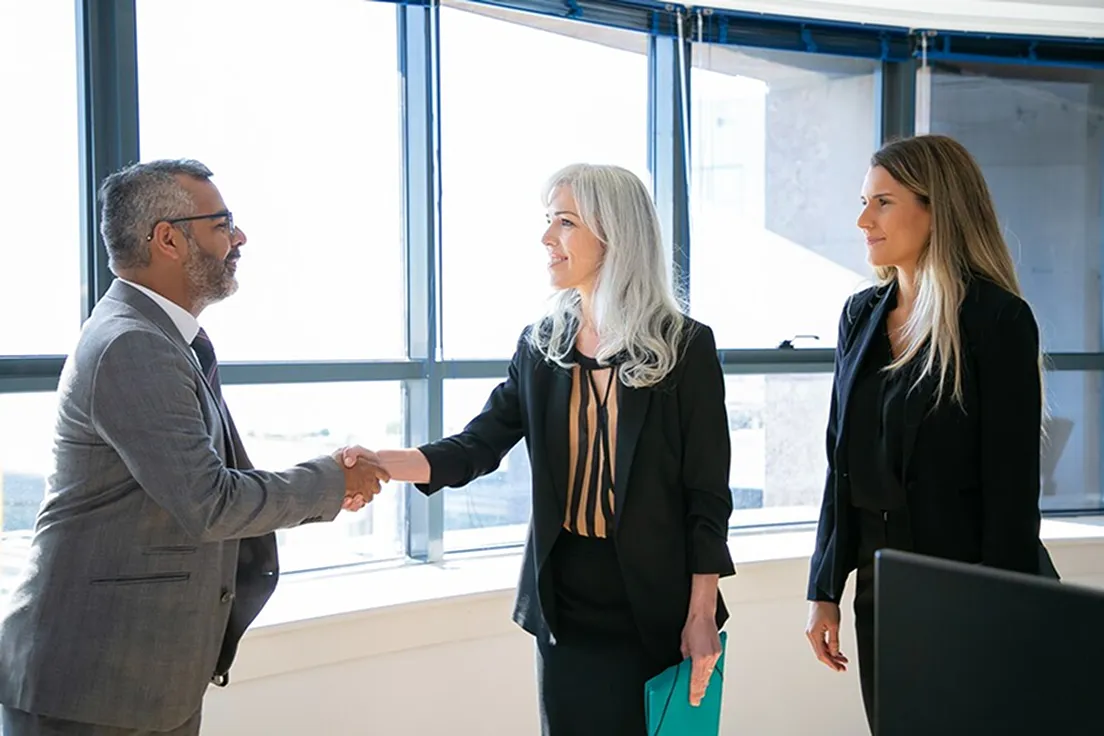 Two women and a man shaking hands in front of a window.