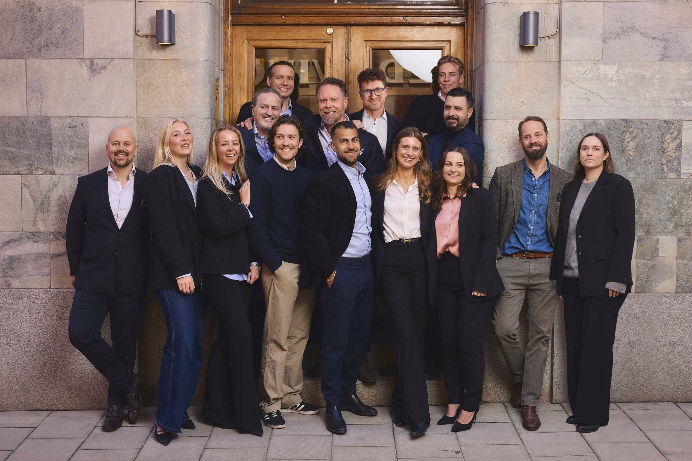 Gruppfoto på femton personer i affärskläder poserande framför en träport och stenvägg.
