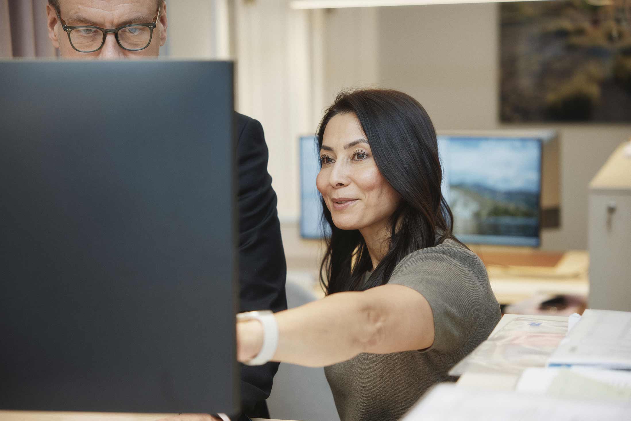 En kvinna pekar på en datorskärm medan en man med glasögon tittar på, båda i ett kontorsmiljö.