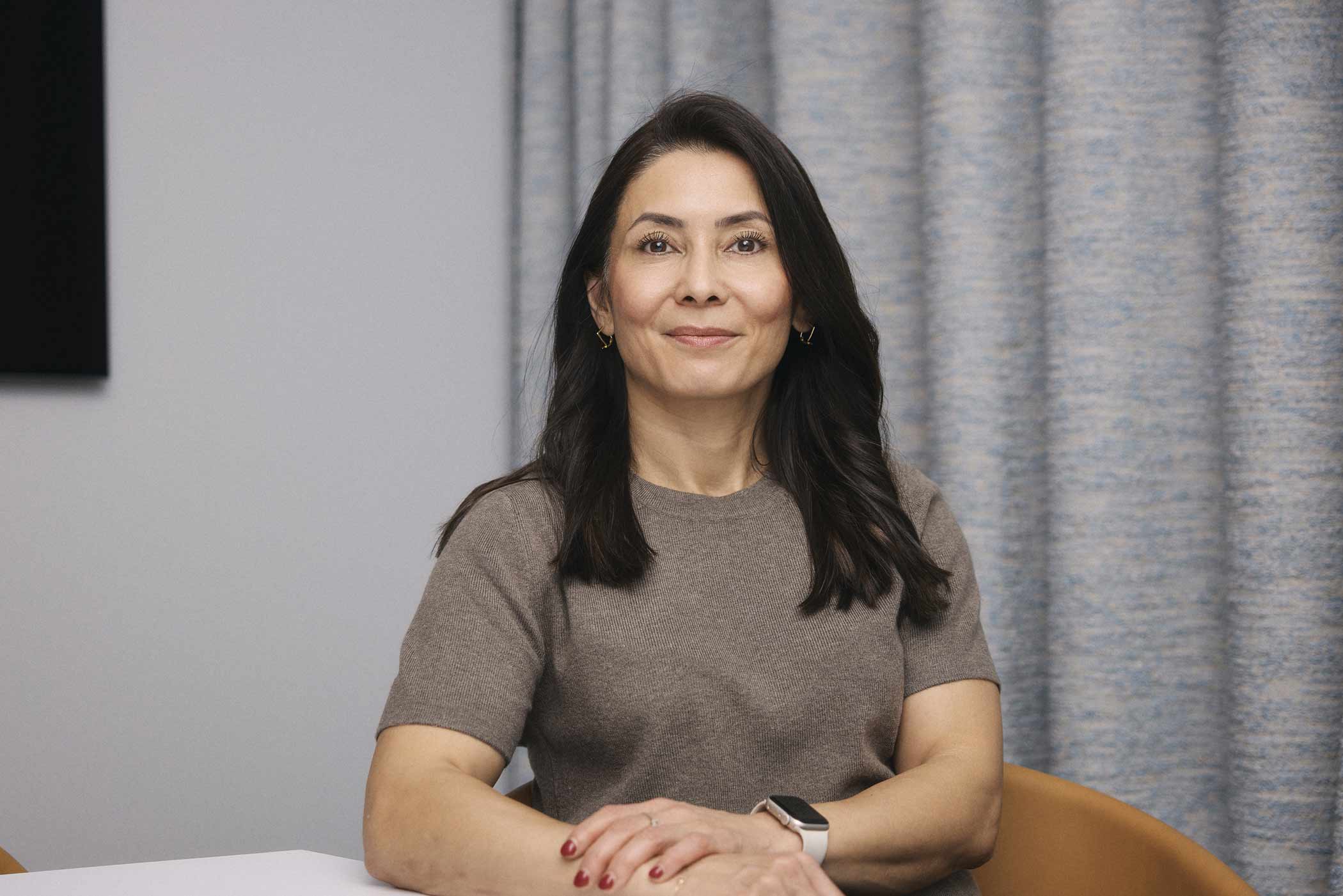 Kvinna med mörkt hår och brun tröja sitter vid ett skrivbord med armarna korsade och ler lätt.