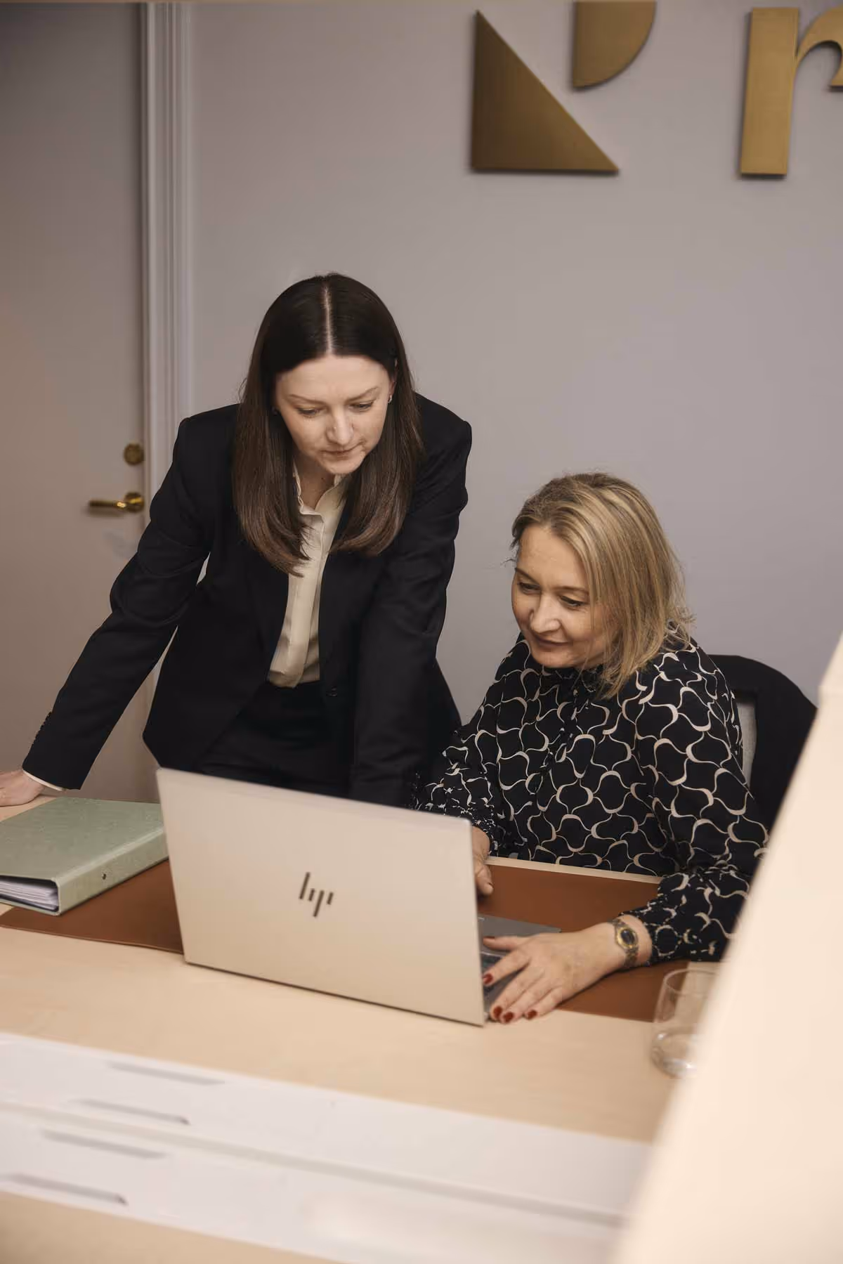 Två kvinnor, en stående i svart kostym och en sittande i mönstrad blus, tittar tillsammans på en laptop på ett kontorsbord.