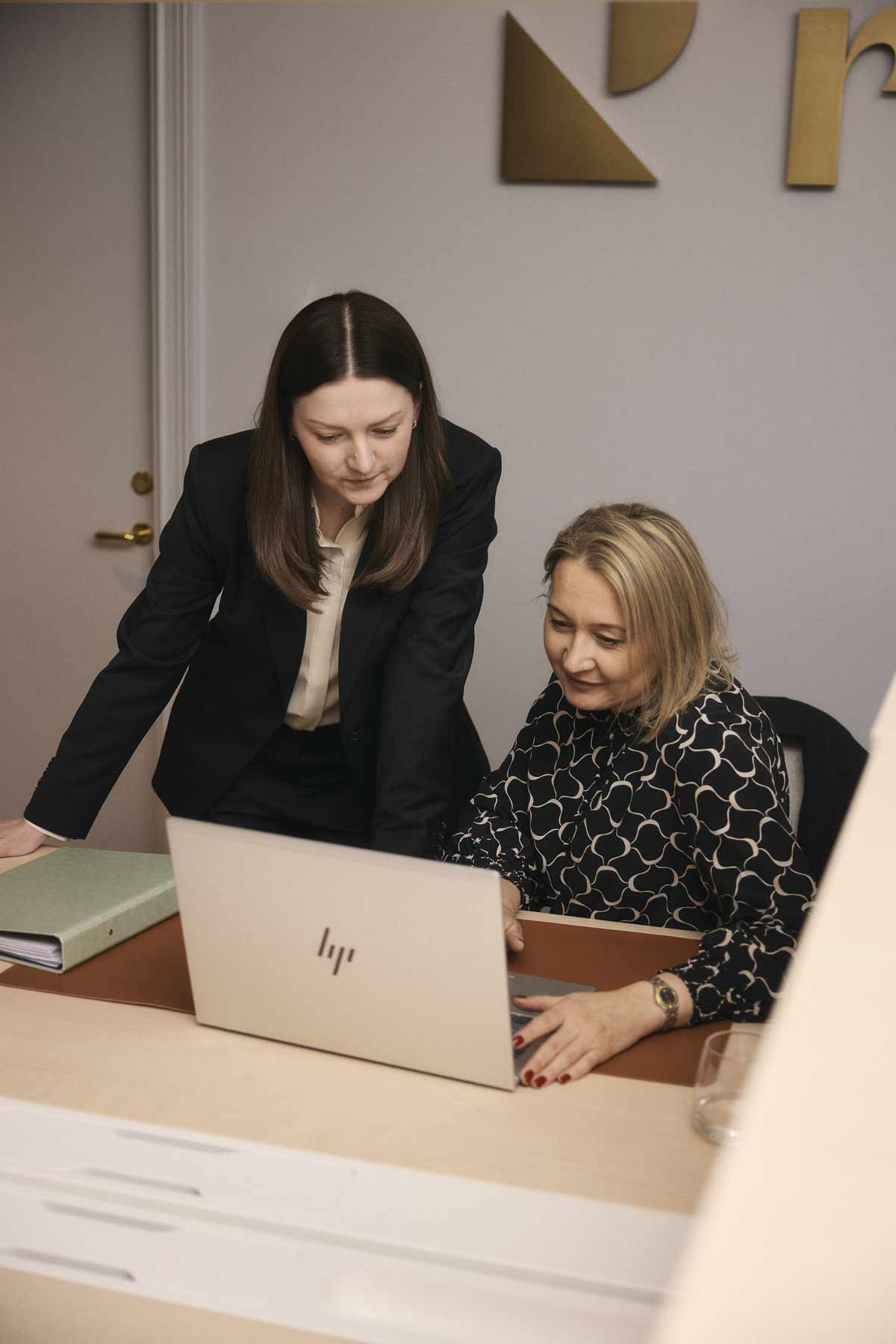 Två kvinnor, en stående i svart kostym och en sittande i mönstrad blus, tittar tillsammans på en laptop på ett kontorsbord.