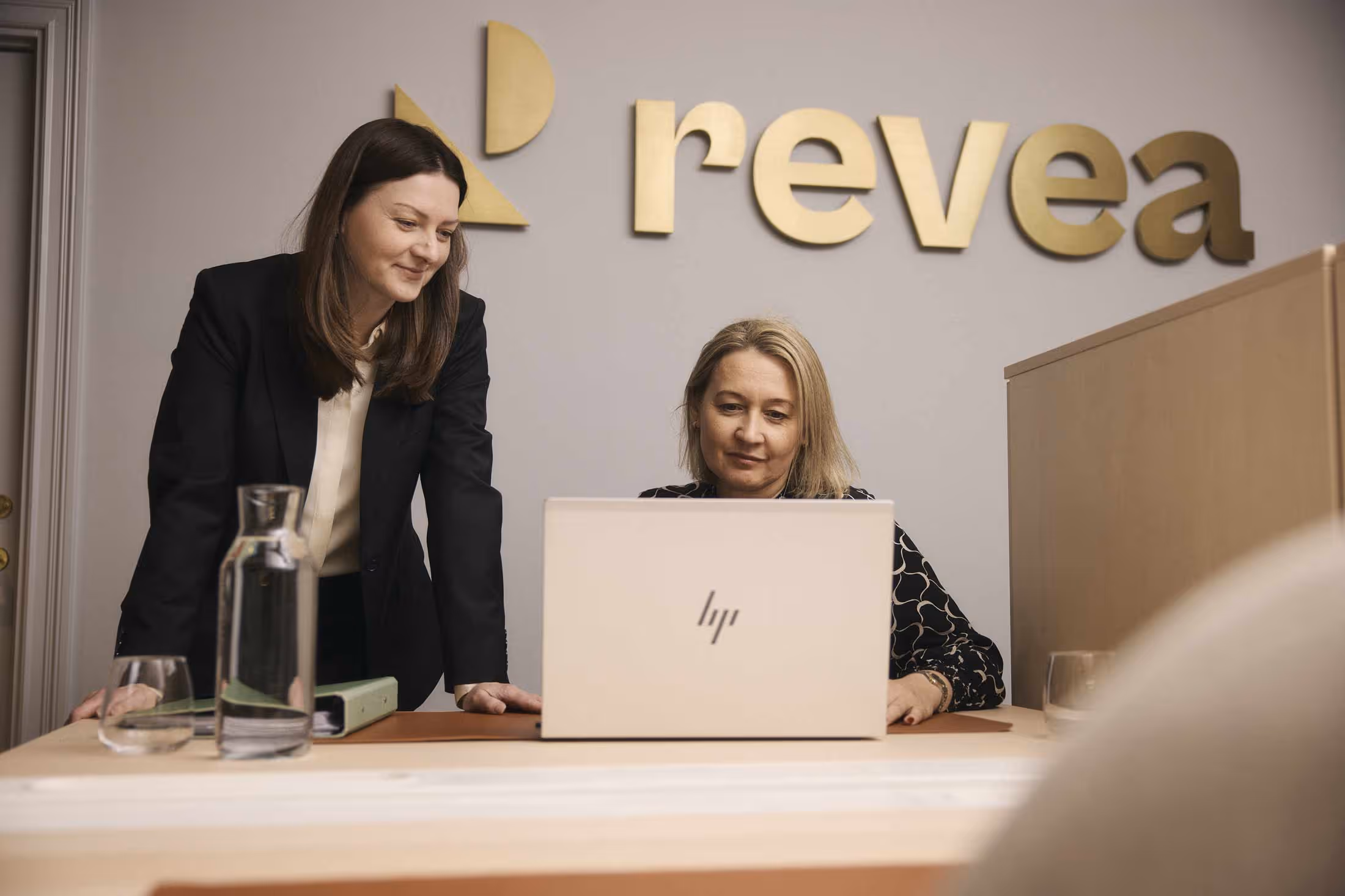 Två kvinnor arbetar tillsammans vid en laptop på ett kontor med 'rekea' logotyp på väggen.