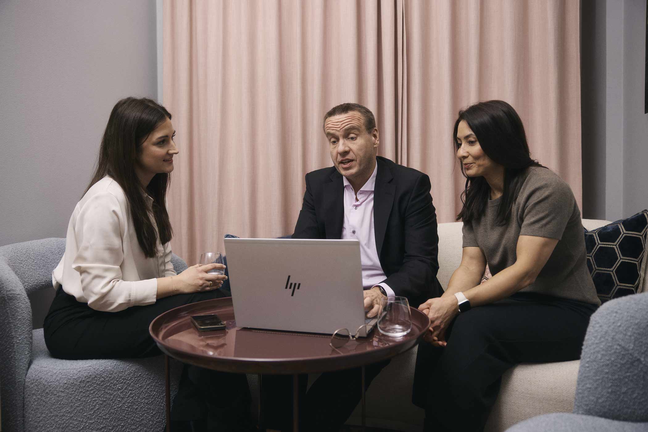 Tre personer, två kvinnor och en man, sitter runt ett bord med en öppen laptop och diskuterar i ett kontorsrum.