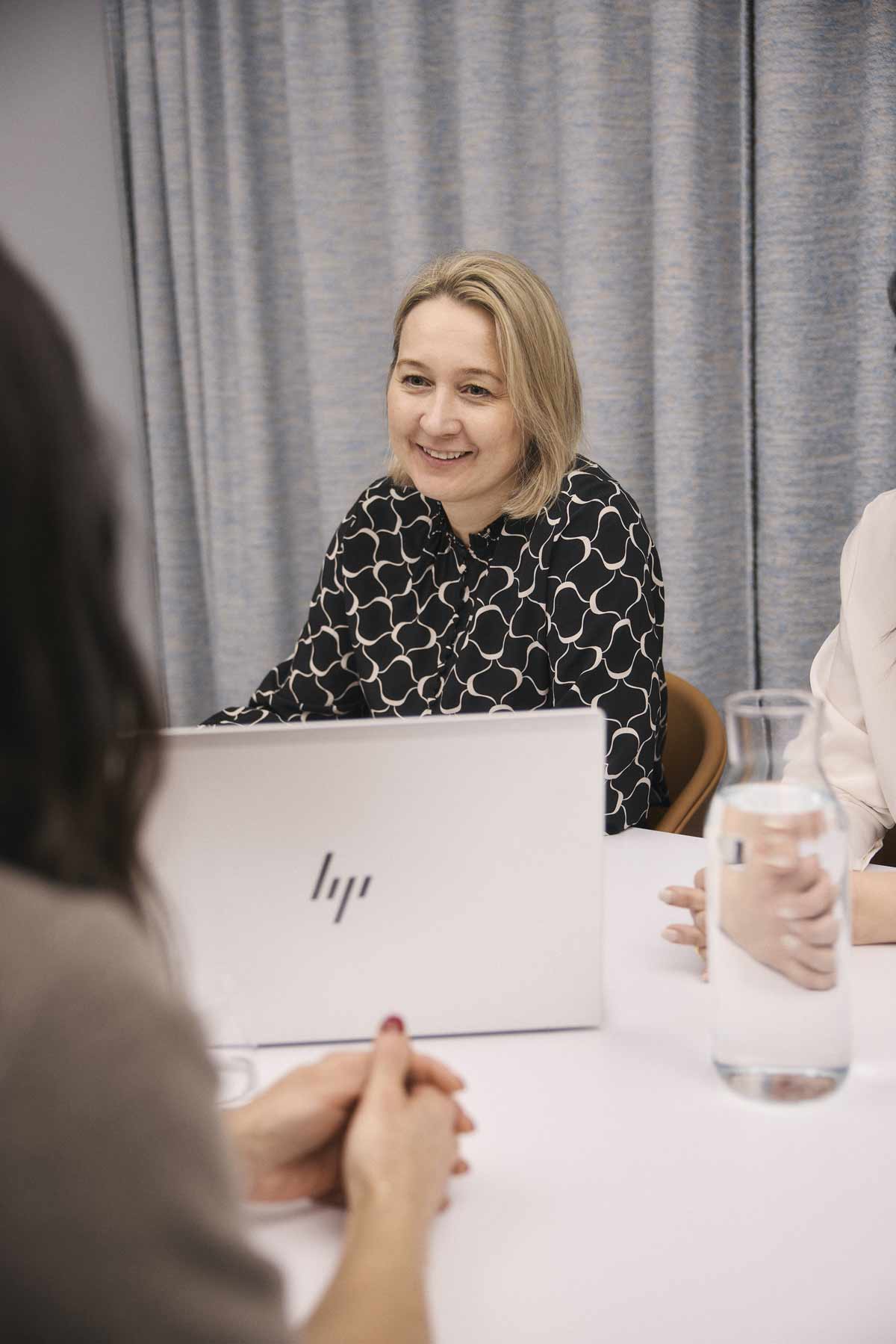 Kvinna som ler och pratar med en annan person över ett bord med en laptop och en karaff vatten.