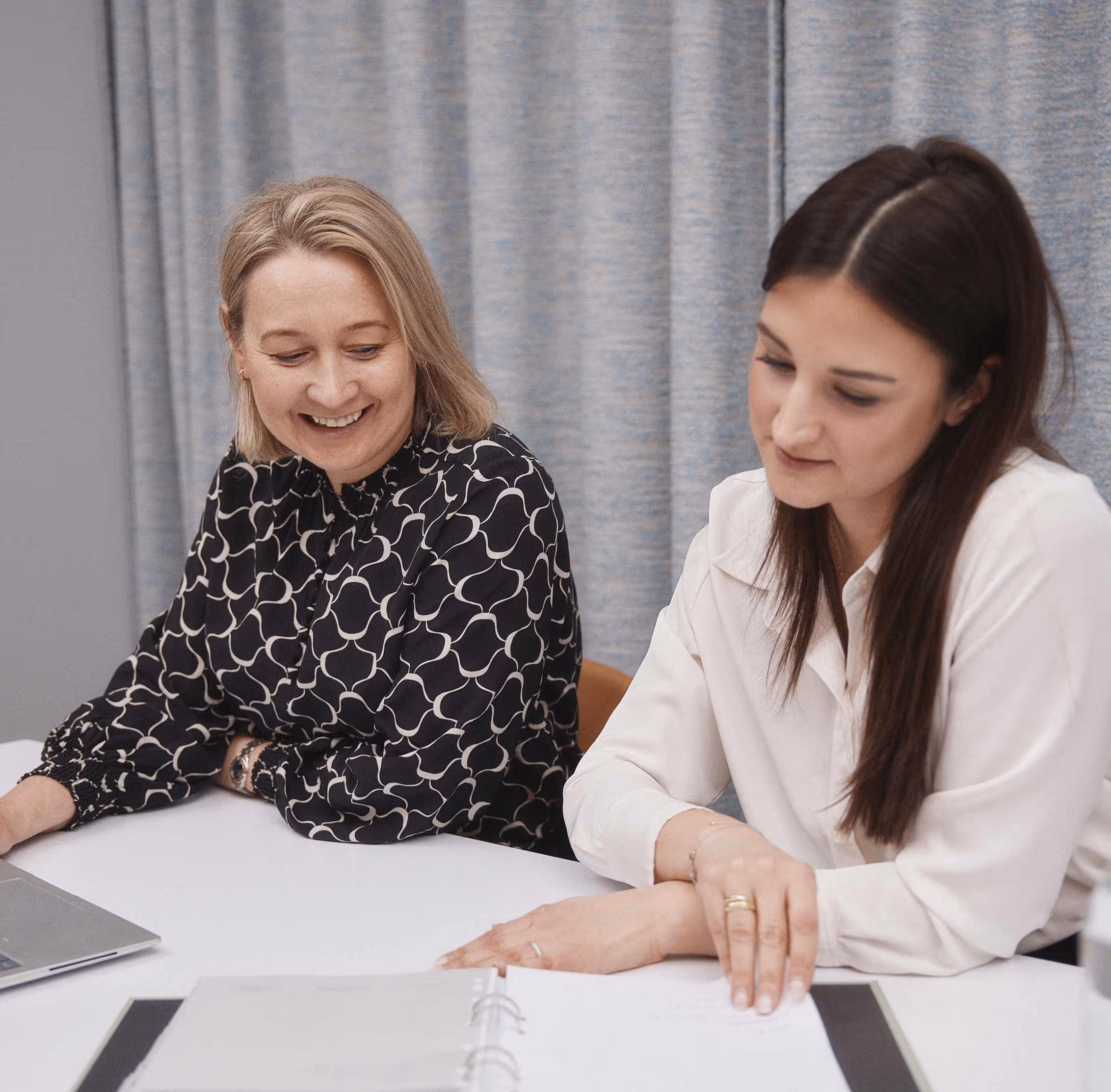 Två kvinnor sitter vid ett bord och ser över dokument i en pärm, med en laptop bredvid.