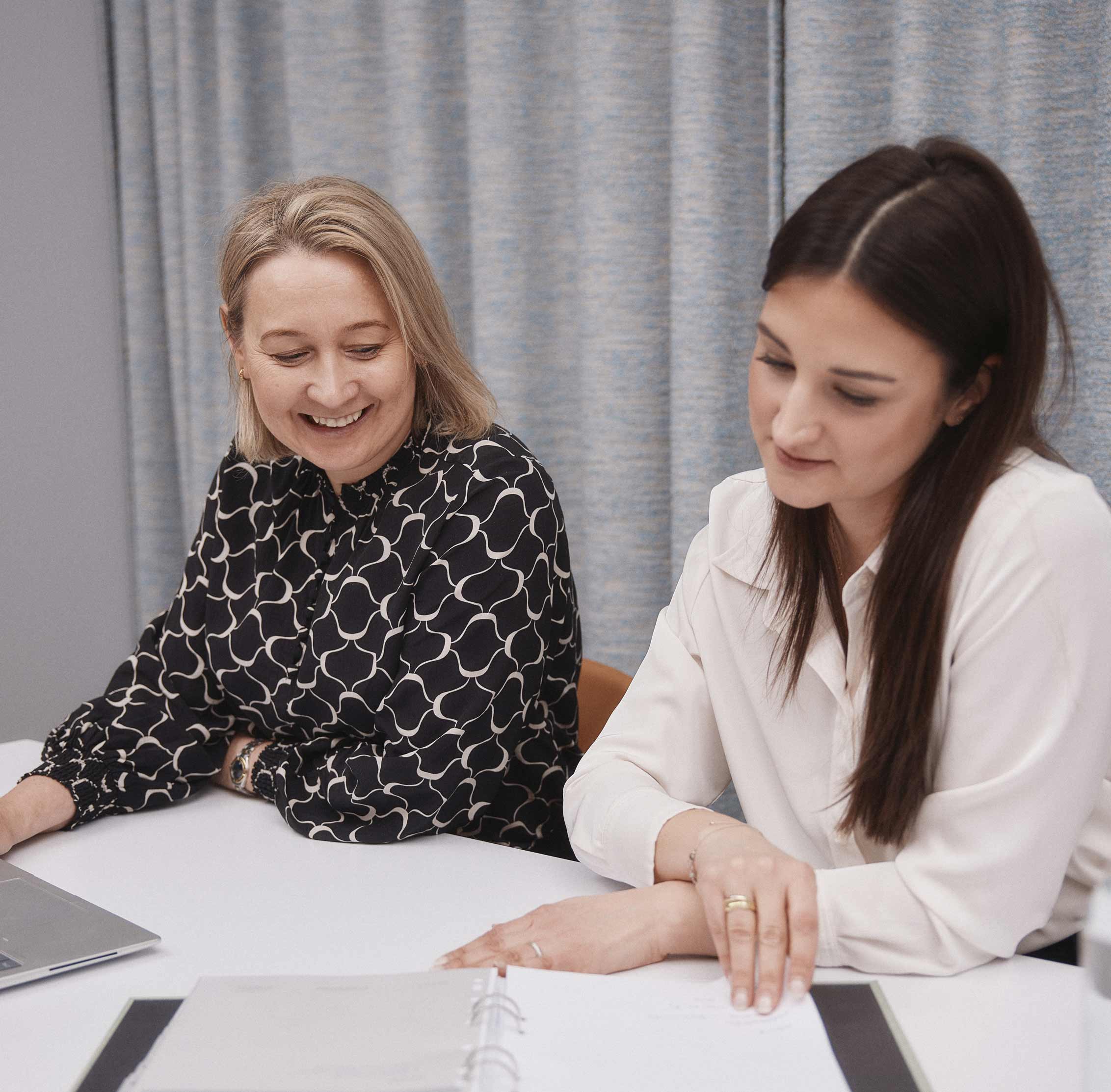 Två kvinnor sitter vid ett bord och ser över dokument i en pärm, med en laptop bredvid.